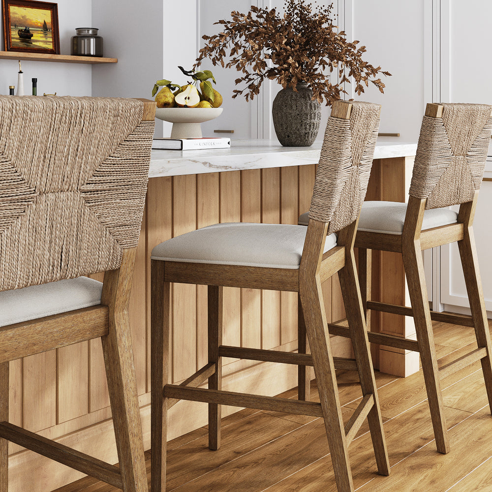 Three wooden bar stools with woven rope backs and light-colored upholstered seats arranged along a marble-topped kitchen island.