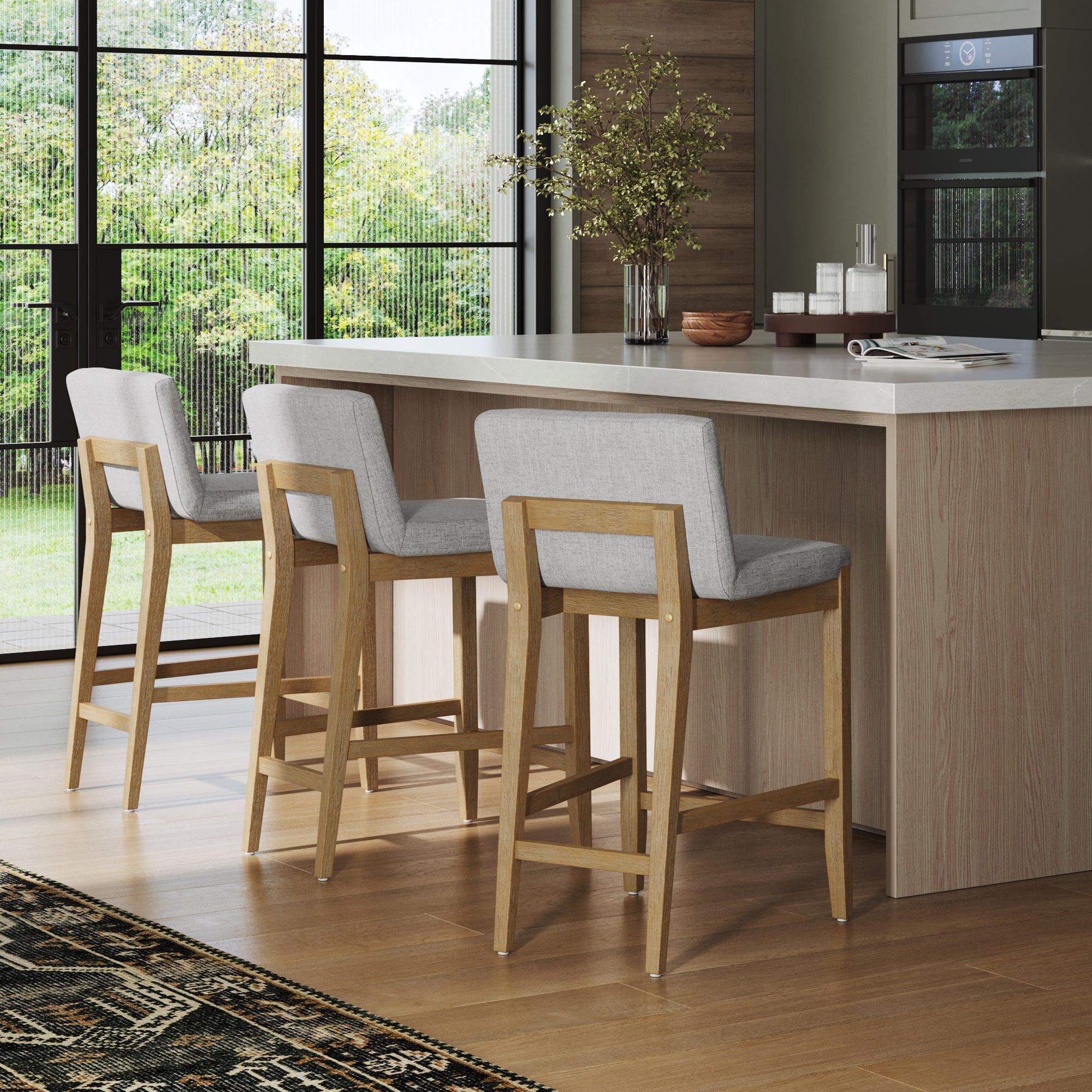 A modern kitchen features a light wood island, three Nathan James Upholstered Counter Height Bar Stools with backs in gray, large windows with greenery views, and a patterned rug on the wooden floor.