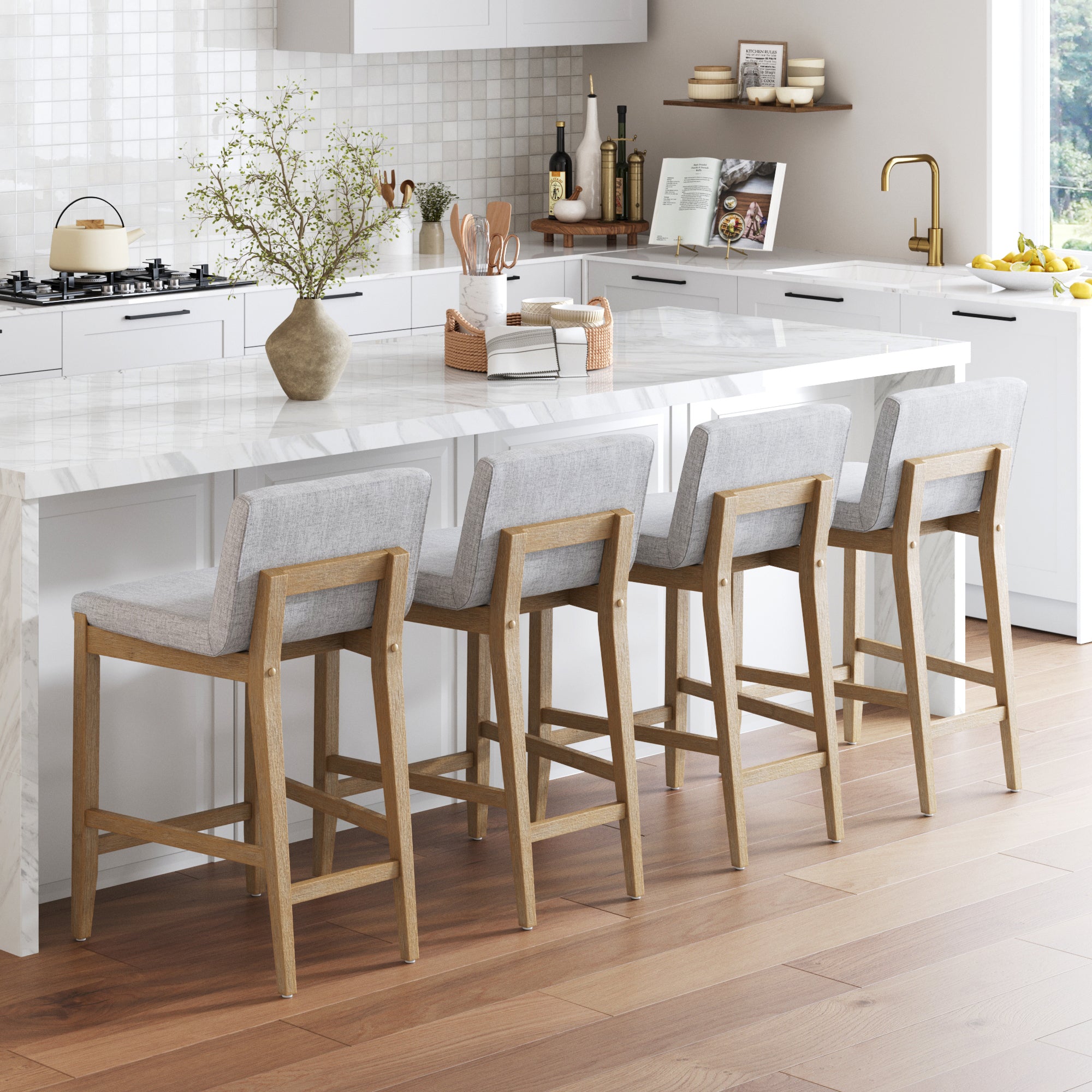 A modern kitchen features a white marble island, four Nathan James Upholstered Counter Height Bar Stools with gray backs and cushions, sleek white cabinets, a vase with branches, cooking essentials on the counter, and sunlight streaming in.