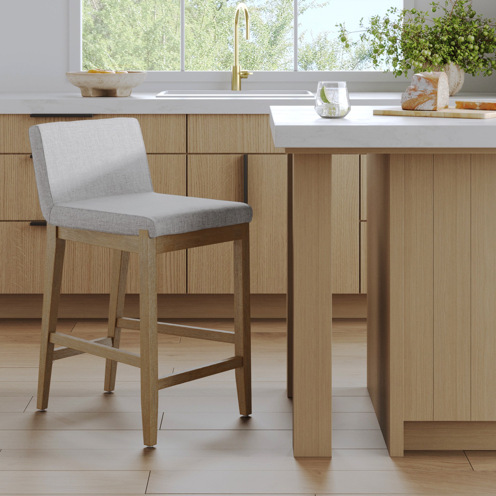 A modern kitchen features light wood cabinets, a white countertop, and the Nathan James Modern Upholstered Counter Height Bar Stool w/ Back Gray. By the window with greenery, a glass of water and bread on a cutting board rest on the counter.