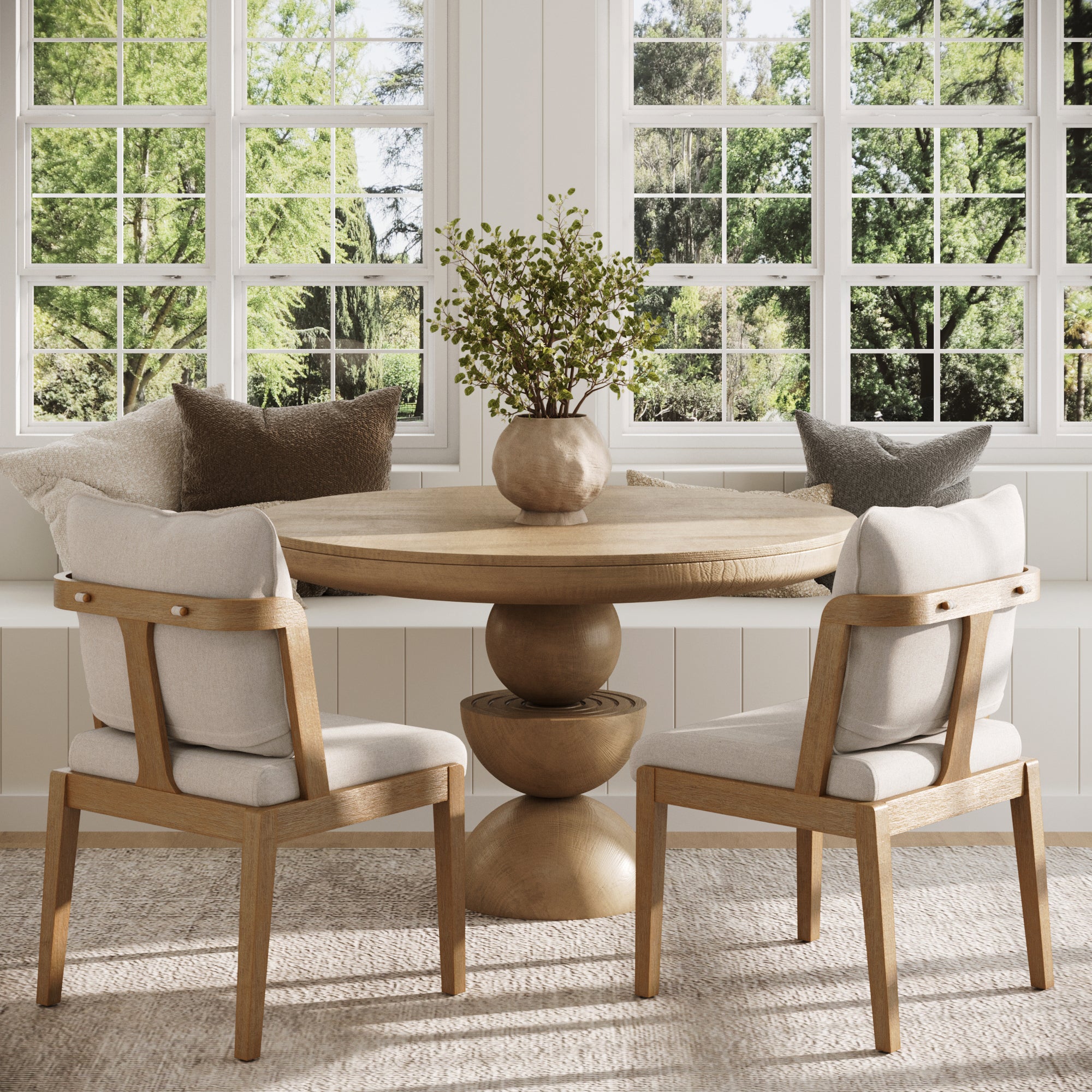 A cozy breakfast nook with a round wooden table, two Nathan James Scandinavian Dining Chairs in light brown, a vase with greenery, window seats with pillows, and large windows overlooking a lush green view.