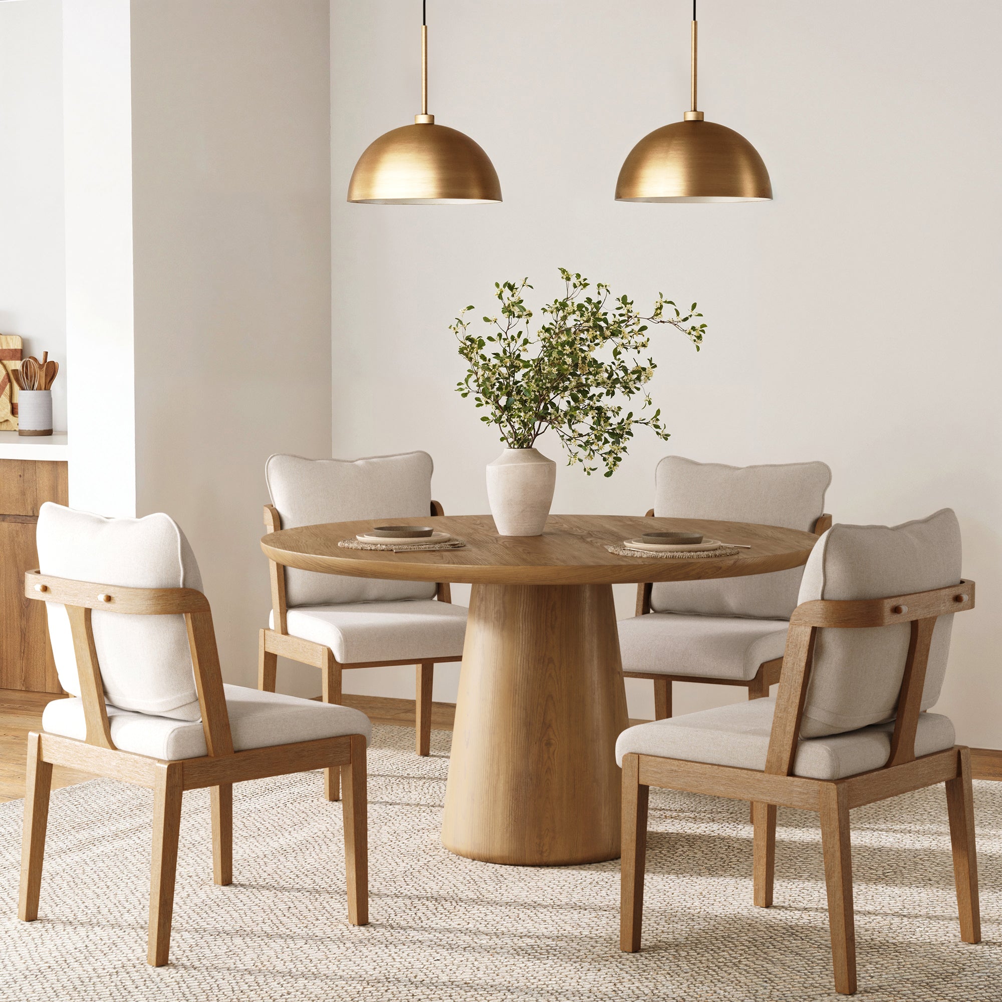 A round wooden dining table with four Nathan James Scandinavian Dining Chairs in light brown, a white vase with green foliage centerpiece, and two brass pendant lights in a modern, neutral-toned dining room.
