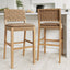 Two Nathan James Faux Leather Woven Bar Stools in brown (set of 2), featuring cushioned seats and backrests, sit before a white kitchen island with snacks and appliances in the background.