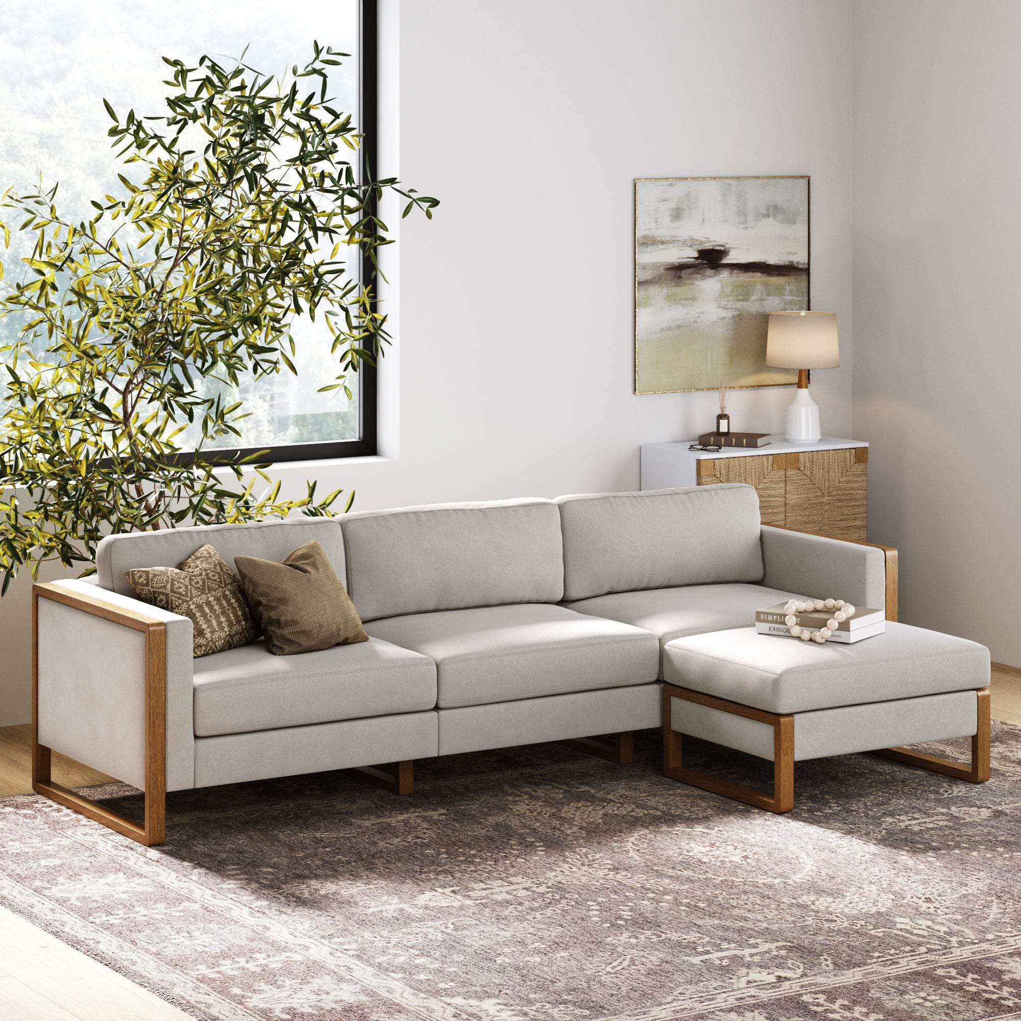 A modern living room with the Nathan James Upholstered Sofa Standard Seat Textured Gray (Modular Sofa Part), plush back cushions, an ottoman, neutral throw pillows, a patterned rug, potted plant by a window, sideboard with lamp, and wall art.