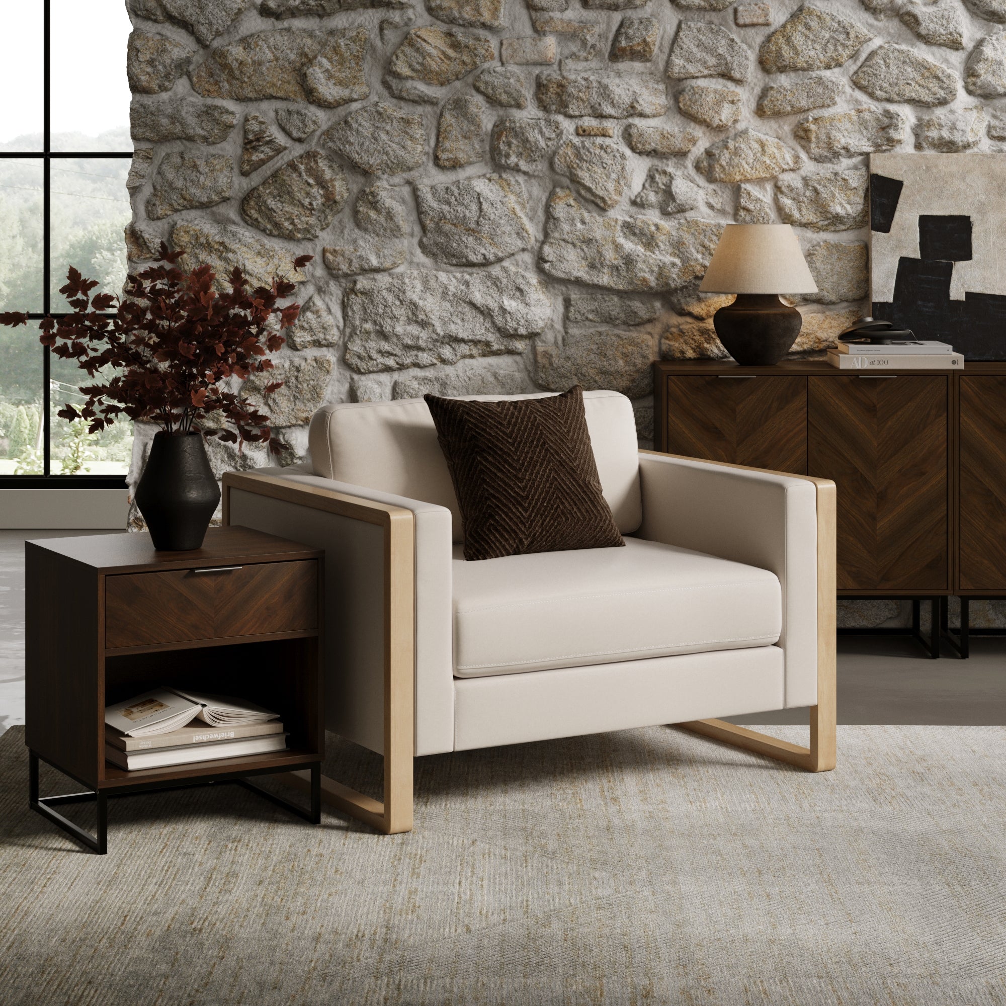 A modern living room features a beige armchair with a dark throw pillow, a wooden side table with a vase of red leaves, a stone wall, and a lamp on a wooden cabinet, creating a cozy, stylish atmosphere.
