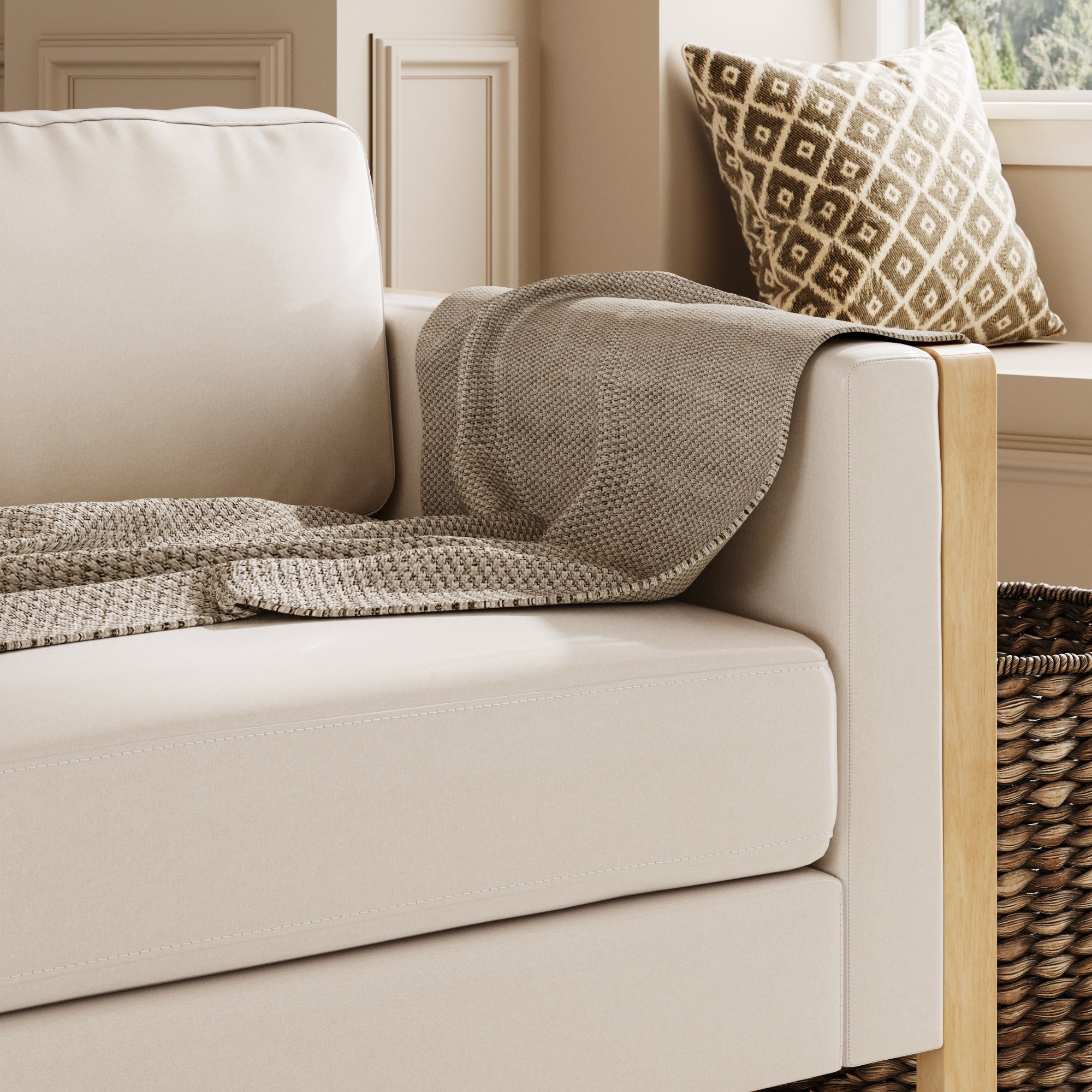 A beige sofa with a textured gray blanket draped over the armrest, a patterned cushion in the corner, and a woven basket next to the sofa, set in a bright, cozy room.