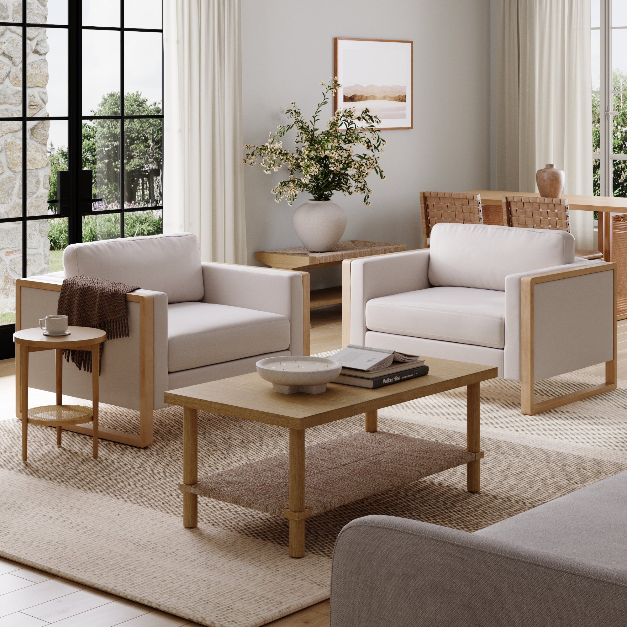 A modern living room with two cream armchairs, a wooden coffee table, a side table with a brown throw, a large vase with branches, light curtains, and large windows letting in natural light.
