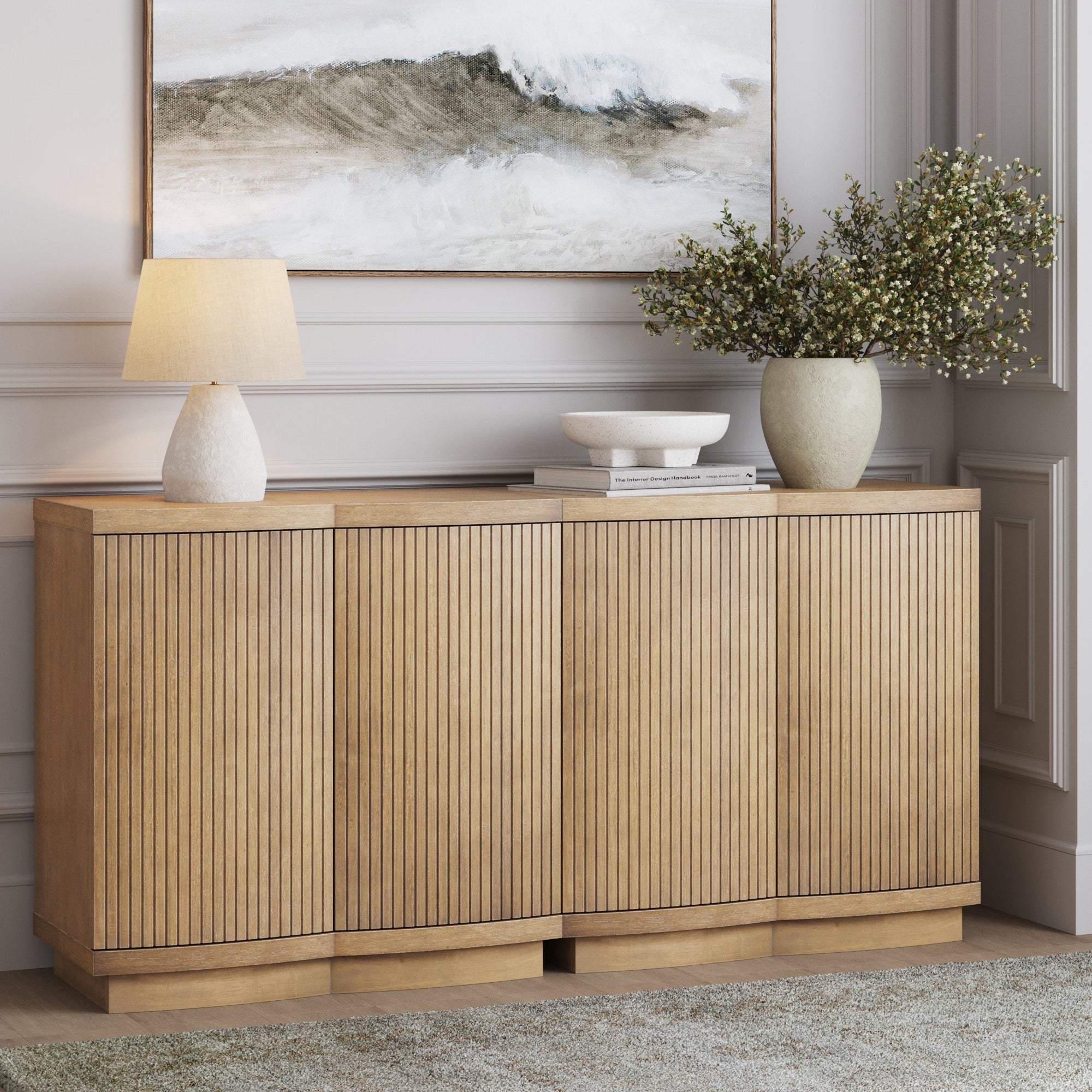 A wooden sideboard with vertical grooves stands against a wall, topped with a lamp, stacked books, a decorative bowl, and a vase with leafy branches. Above hangs a painting of ocean waves in neutral tones.