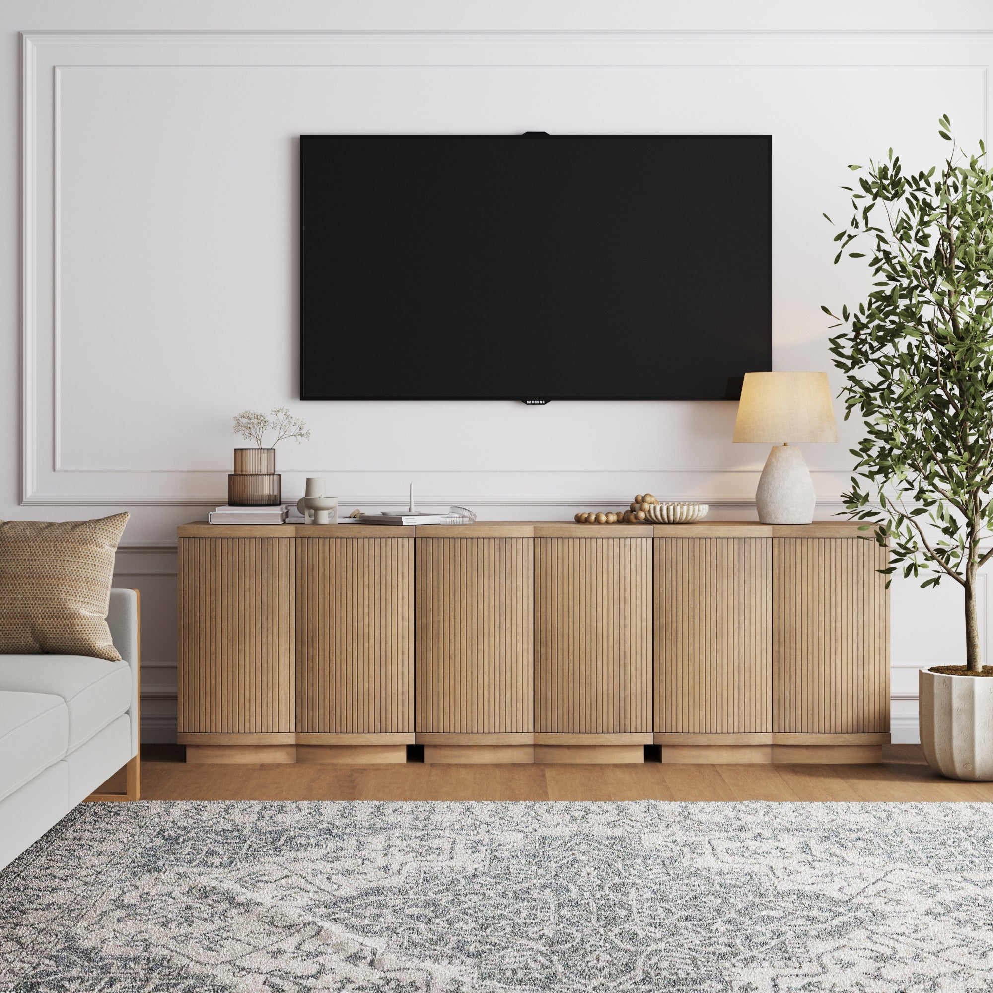 A modern living room features a large flat-screen TV mounted on a white wall above a wooden sideboard. The sideboard holds a lamp, books, and decor. A patterned rug covers the floor and a potted plant sits nearby.