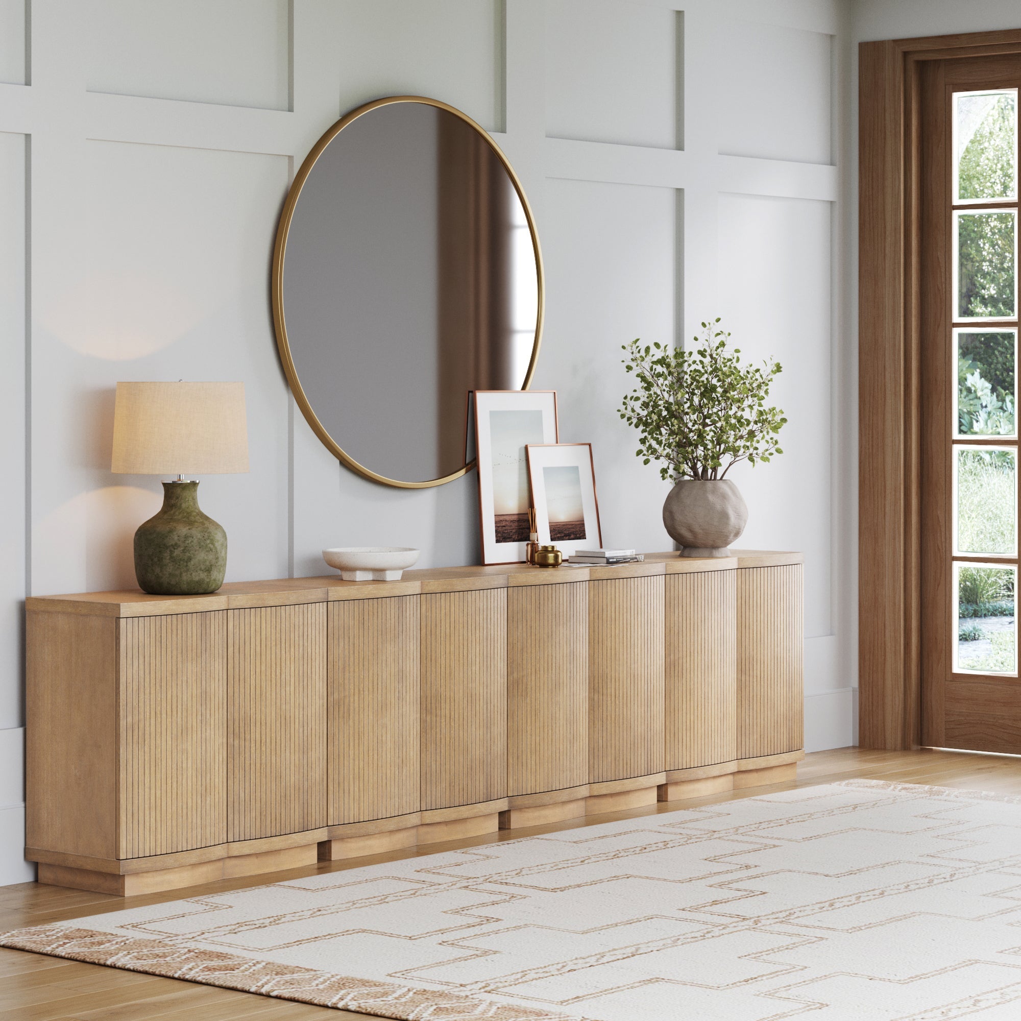 A modern living room features a wooden sideboard with a round mirror above it, a table lamp, framed photos, a decorative bowl, and a vase with greenery. The space has light walls, wood accents, and a patterned rug.