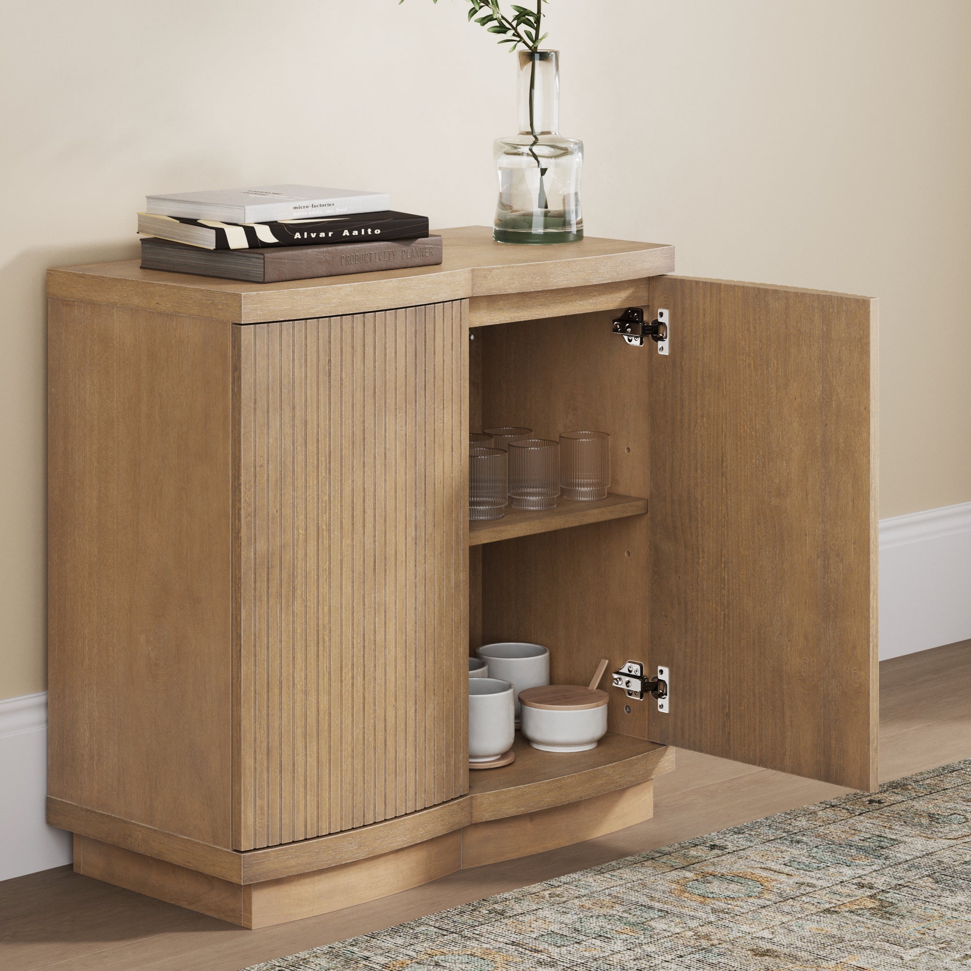 A small wooden cabinet with two doors, one open to reveal cups and bowls inside. The cabinet top holds stacked books and a glass vase with greenery. The cabinet stands against a beige wall on a wooden floor.