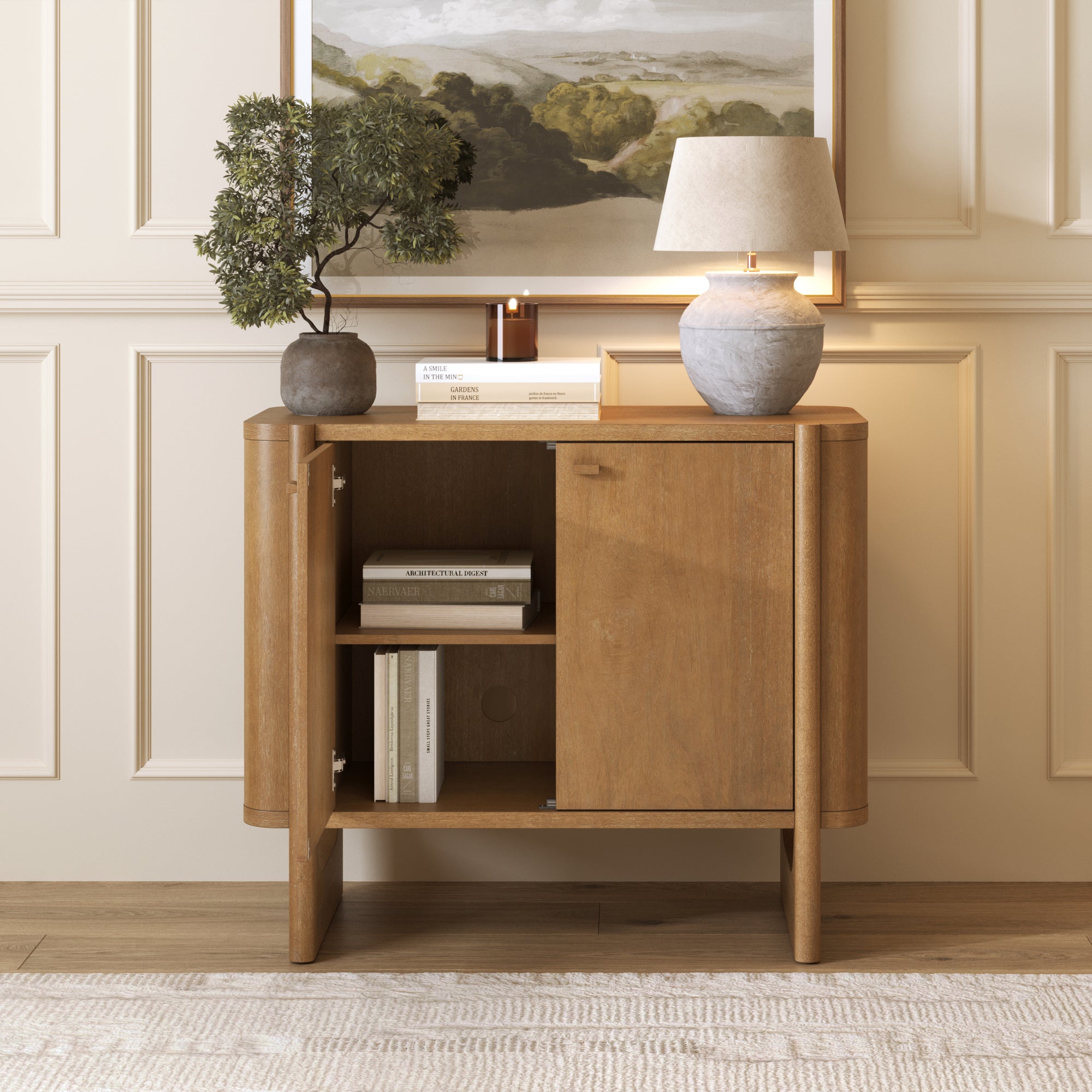 A wooden cabinet with one open shelf and a closed door stands against a paneled wall. On top are a potted plant, books, a lamp, and a framed landscape painting. A light rug covers the floor.