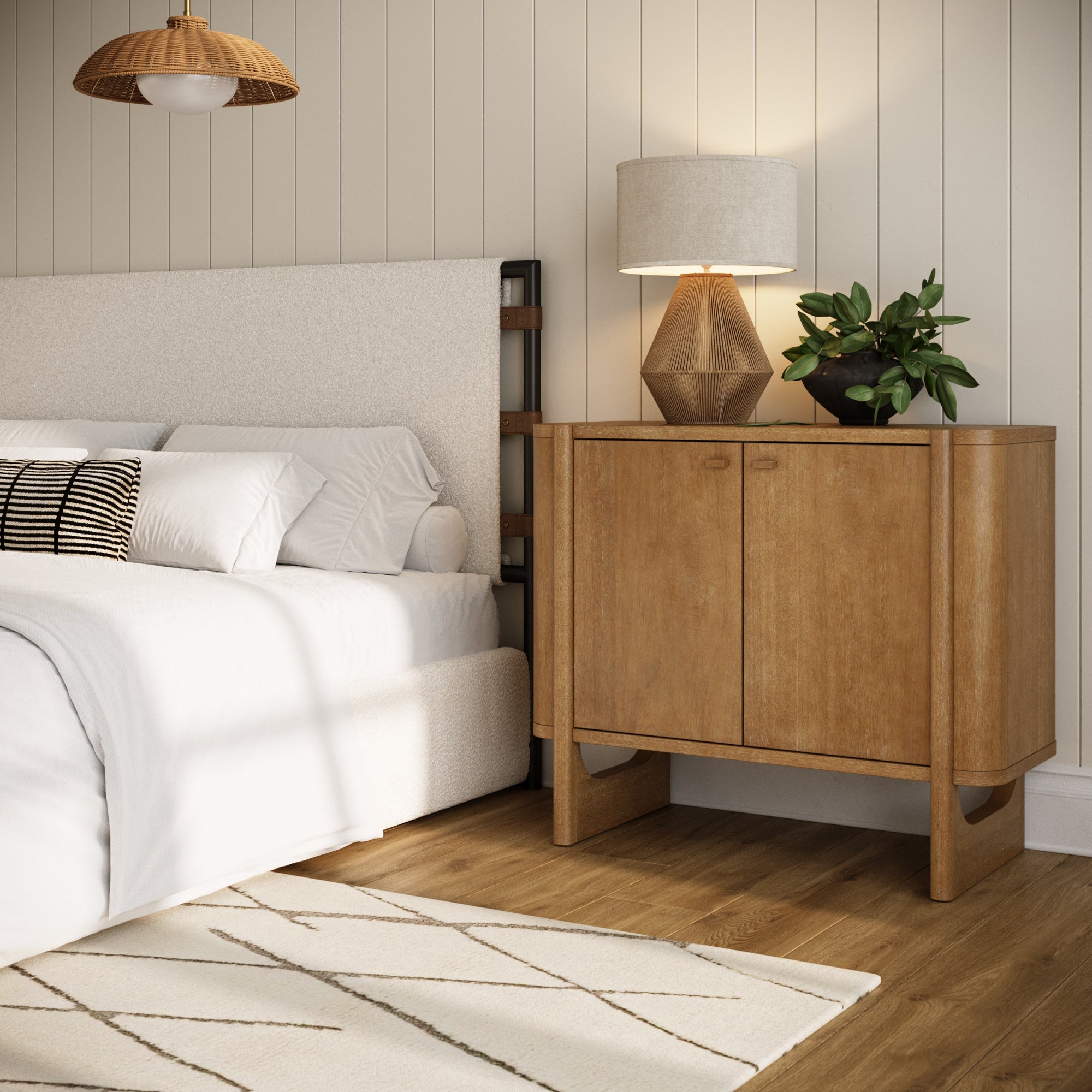 A cozy bedroom with a white bed, wood nightstand, lamp, and plant. The room features light-colored walls, wooden flooring, and a rug with a geometric pattern. A pendant light hangs from the ceiling.