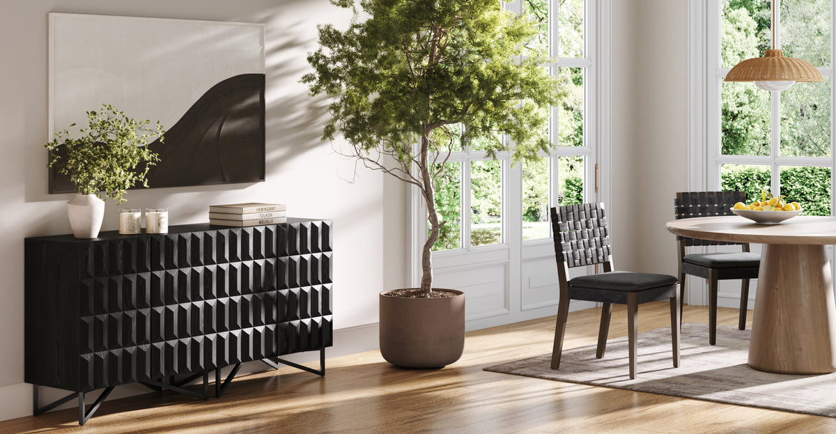 A black textured sideboard topped with a plant, candles, and books, standing next to a potted tree and a wooden dining set.