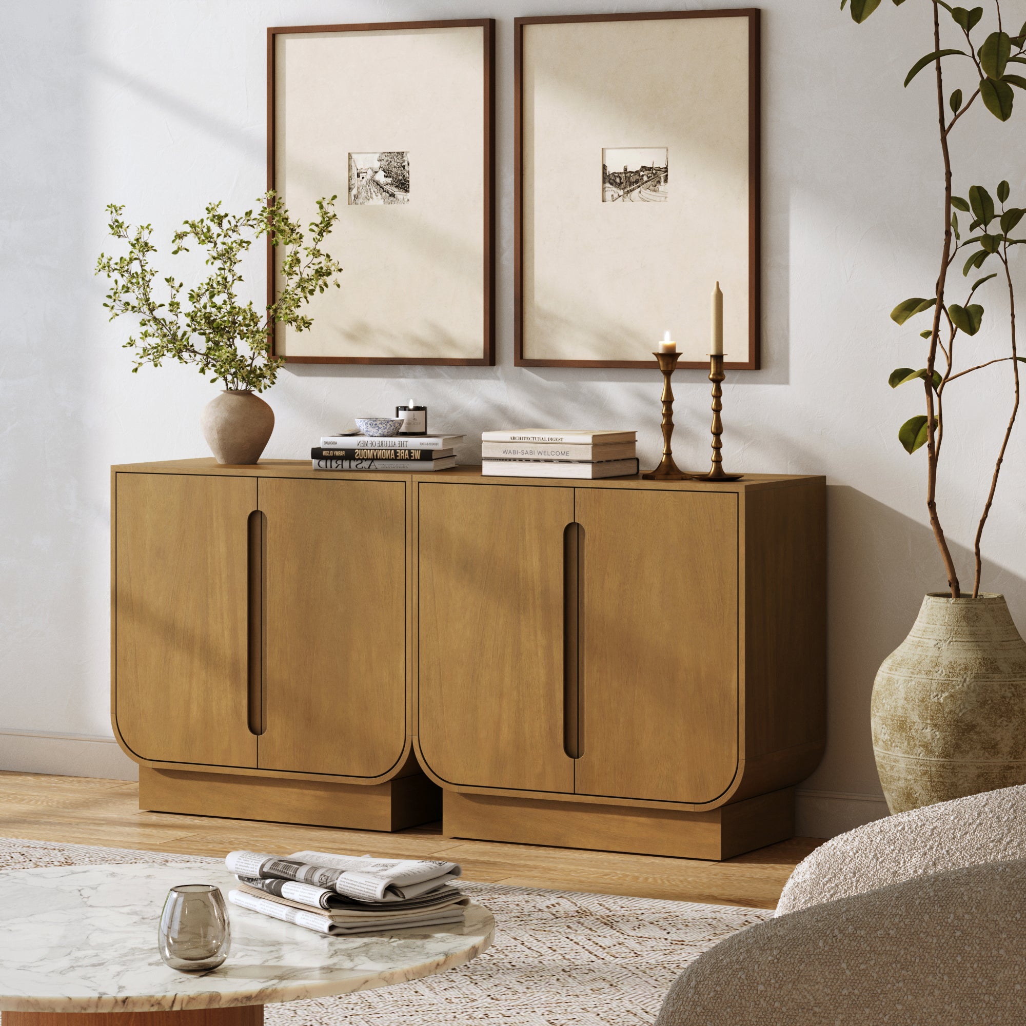 A modern living room features the Nathan James Veneer Arched Accent Cabinet or TV Stand (Set of 2) in light brown, stacked with books and decor. Two framed artworks hang above, alongside a round marble coffee table and a neutral armchair.