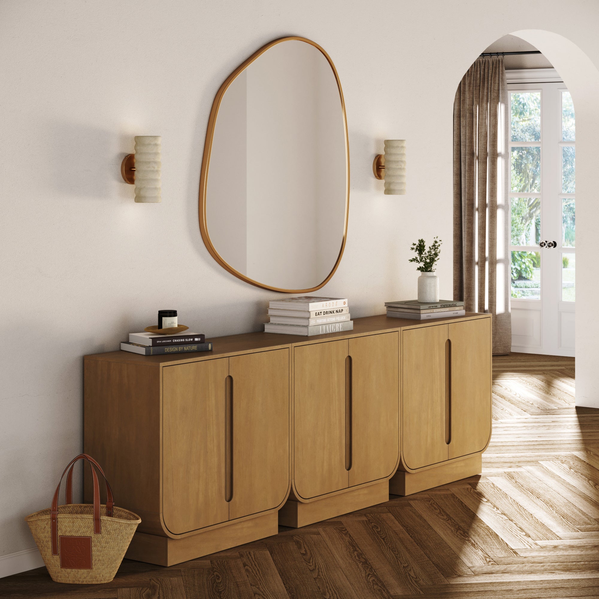 A modern entryway features a wavy-edged mirror above the Nathan James Veneer Arched Accent Cabinet or TV Stand Light Brown (Set of 3), styled with books, decor, two wall sconces, a woven bag on the floor, and sunlight streaming through the window.