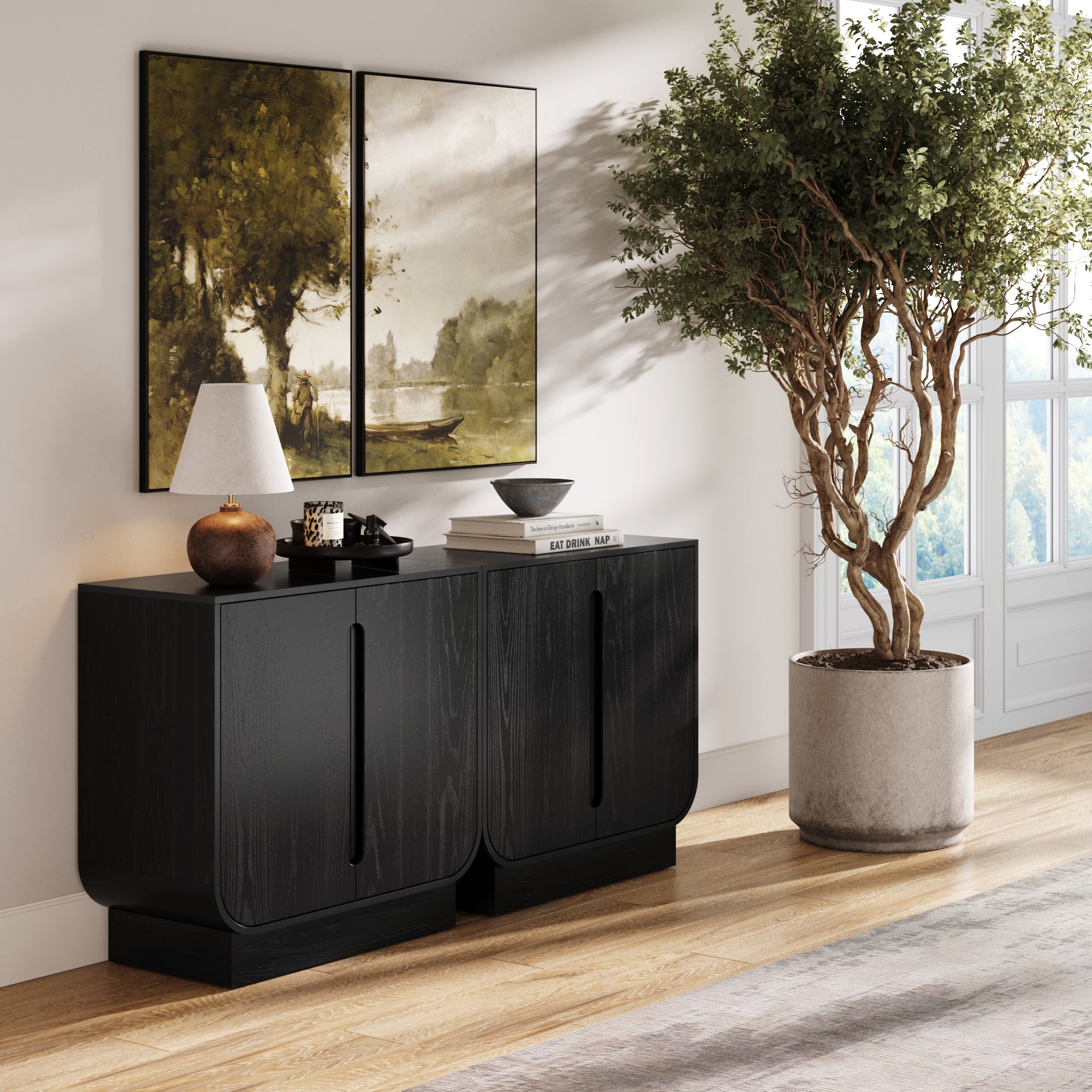 The Nathan James Veneer Arched Accent Cabinet or TV Stand in Black Oak (Set of 2) sits against a white wall beneath two landscape paintings, styled with a lamp, cups, books, and a bowl. A large potted tree stands by the window in sunlight.