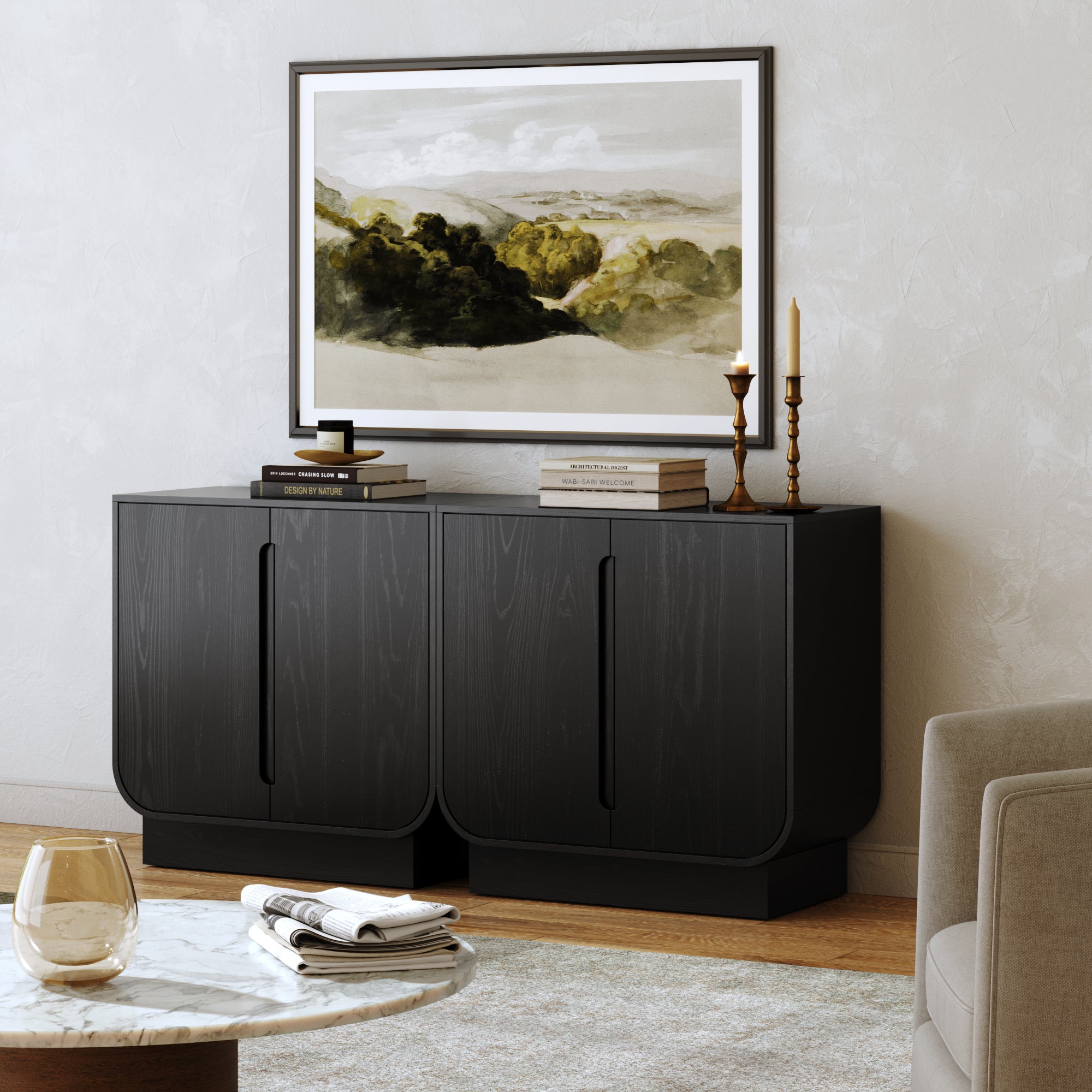 The Nathan James Veneer Arched Accent Cabinet or TV Stand in Black Oak (Set of 2) features curved edges and stands against a light wall, topped with books, gold candlesticks, and framed art. A glass and beige armchair sits nearby.