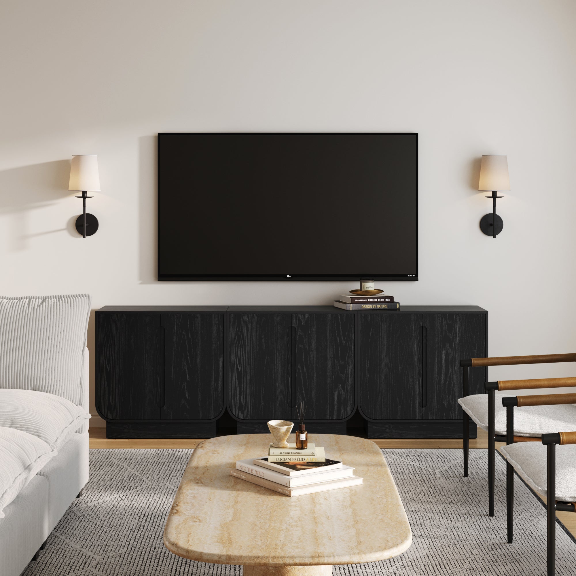 A modern living room with a wall-mounted TV above the Nathan James Veneer Arched Accent Cabinet or TV Stand Black Oak (Set of 3), a beige marble coffee table, a light sofa, a white armchair with black and gold accents, and two wall sconces flanking the TV.