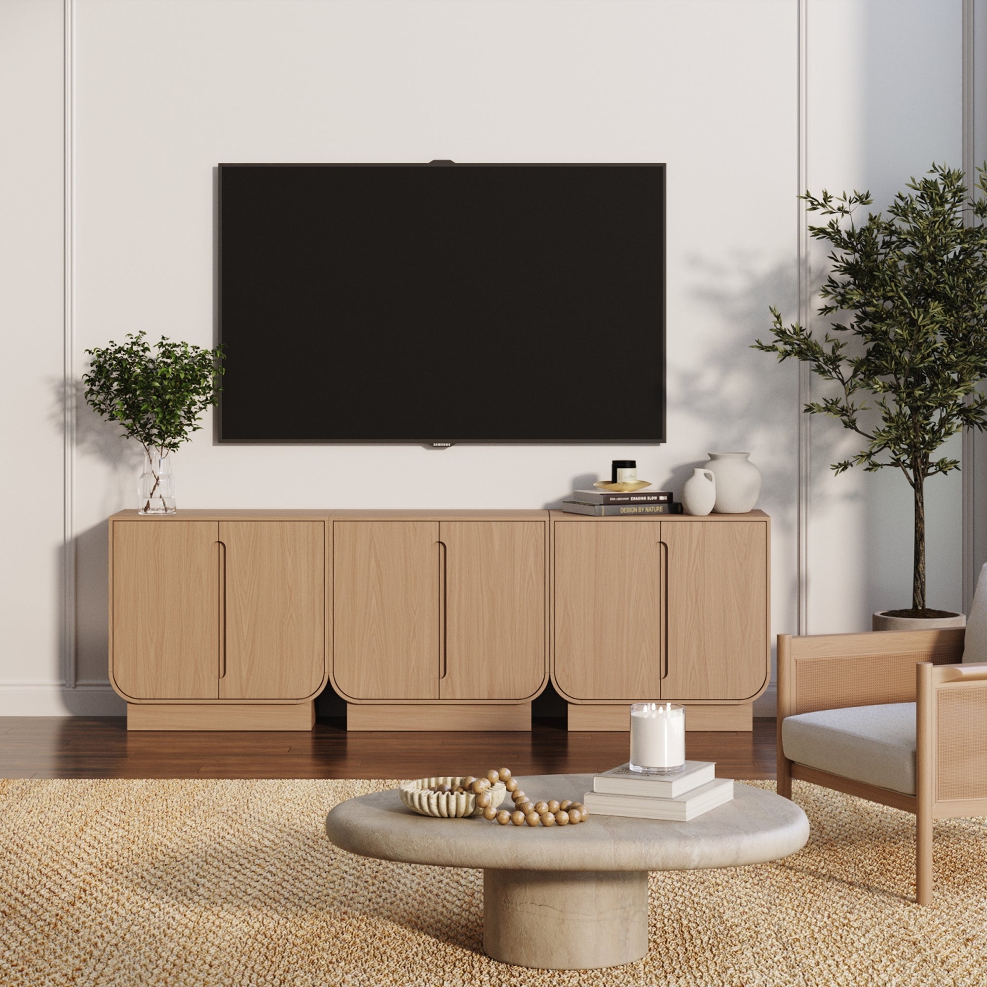 A modern living room with a wall-mounted flat-screen TV above the Nathan James Veneer Arched Accent Cabinet or TV Stand in Raw Oak (Set of 3), a round coffee table, beige armchair, potted plants, and a woven rug on wooden floors.