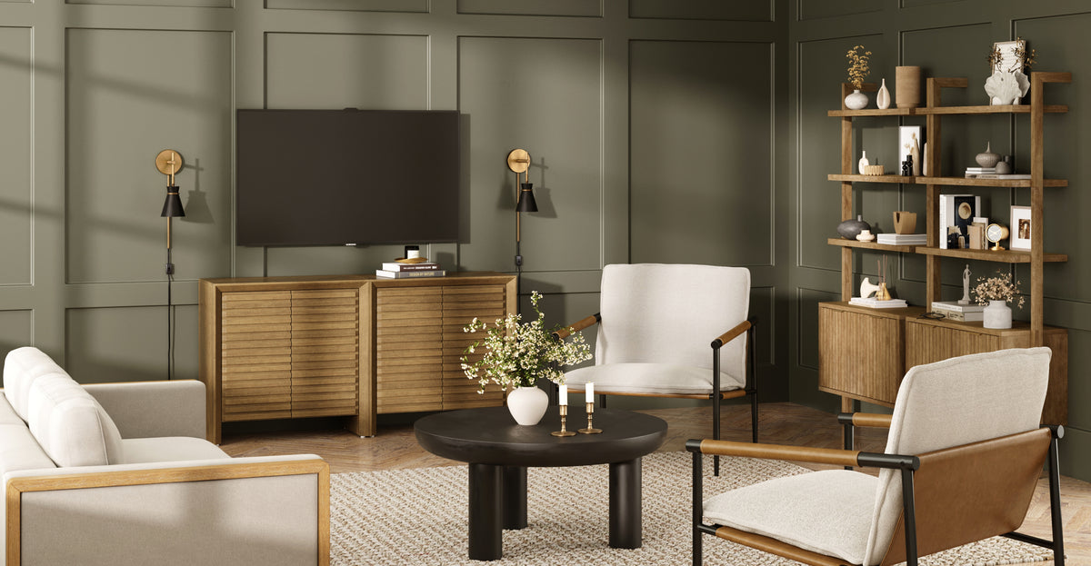 A living room featuring a flat-screen TV mounted above a wooden console, a black coffee table with a flower vase, two cream-colored armchairs, and a wooden open shelving unit against a sage green paneled wall.