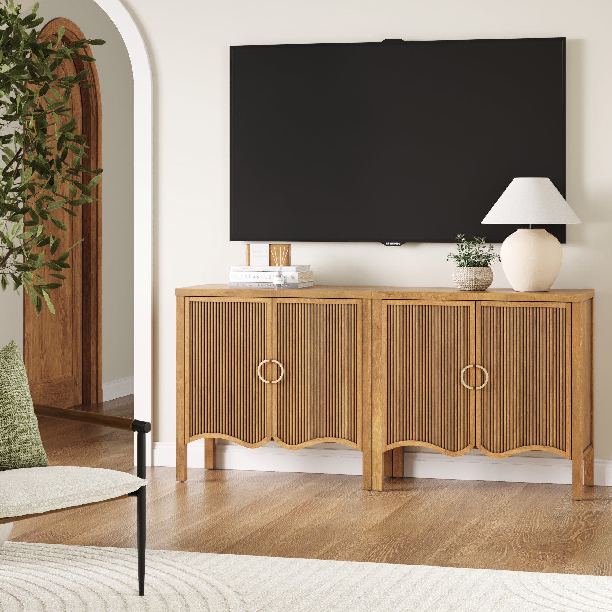 A modern living room features the Nathan James Transitional Fluted Cabinet or TV Stand Light Brown (Set of 2) with fluted doors, solid wood legs, a lamp, plant, books, and a bowl beneath a wall-mounted TV. Light walls and wood flooring complete the space.