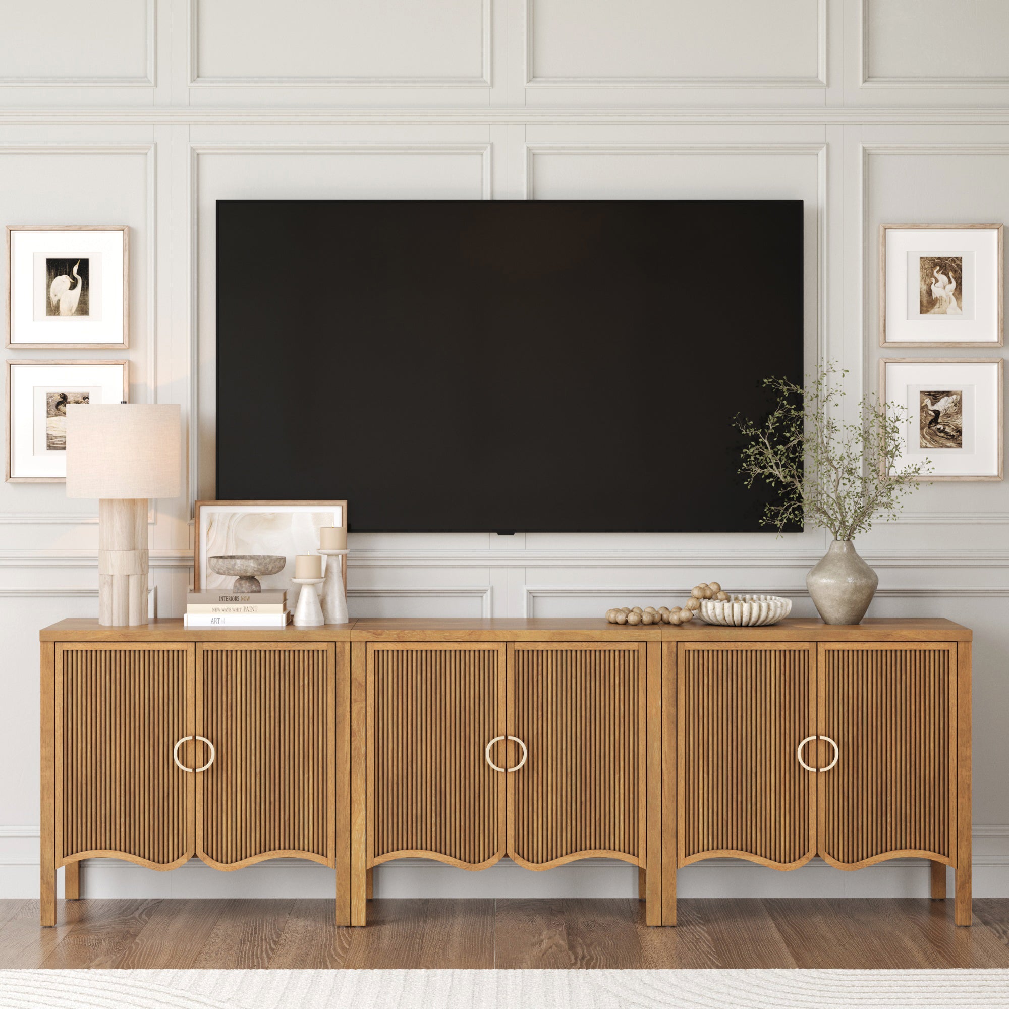 The Nathan James Transitional Fluted Cabinet or TV Stand in light brown (set of 3) features fluted doors and sits beneath a large TV, decorated with a lamp, vases, framed art, and photos against a paneled wall for an elegant touch.
