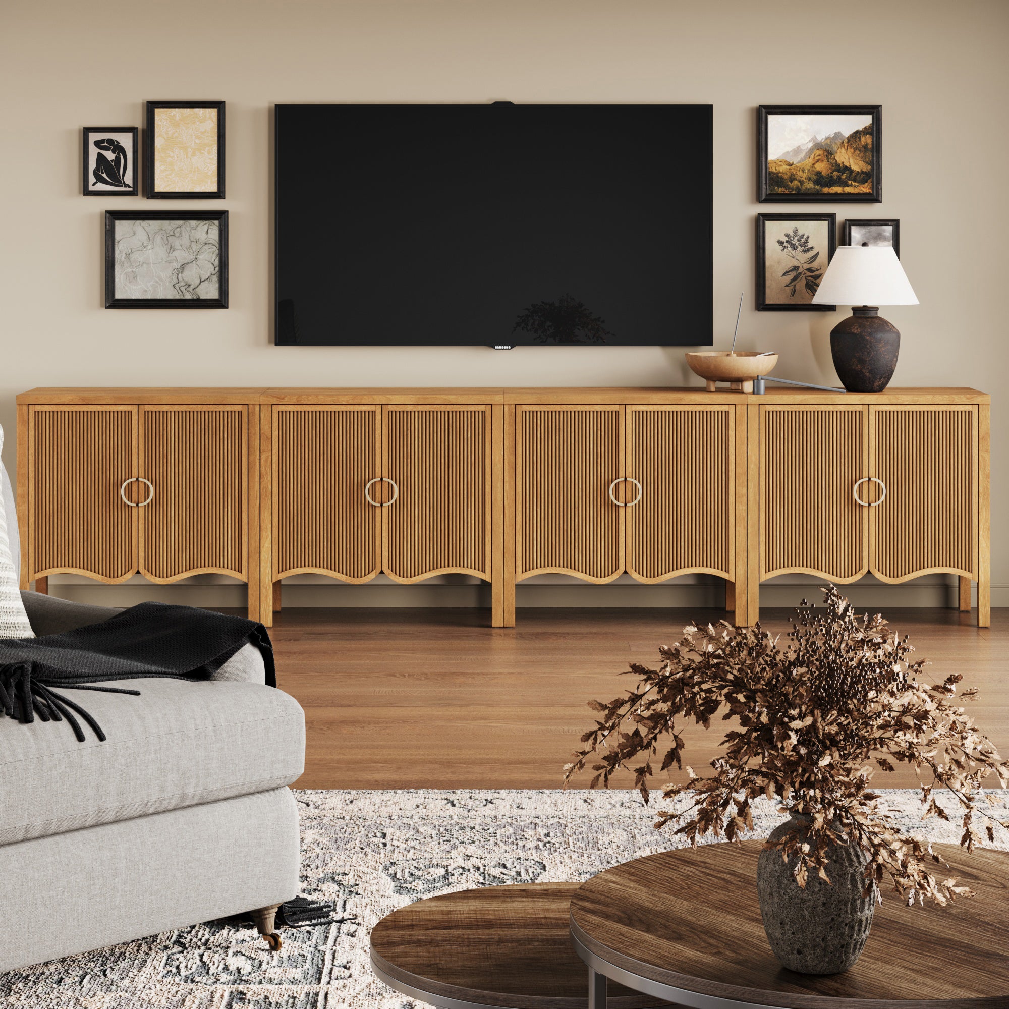 A modern living room features the Nathan James Transitional Fluted Large Credenza Cabinet Light Brown (Set of 4) beneath a wall-mounted TV with framed art. A table lamp, decorative bowl, and vase sit on the credenza; a beige sofa and round coffee table are in front.