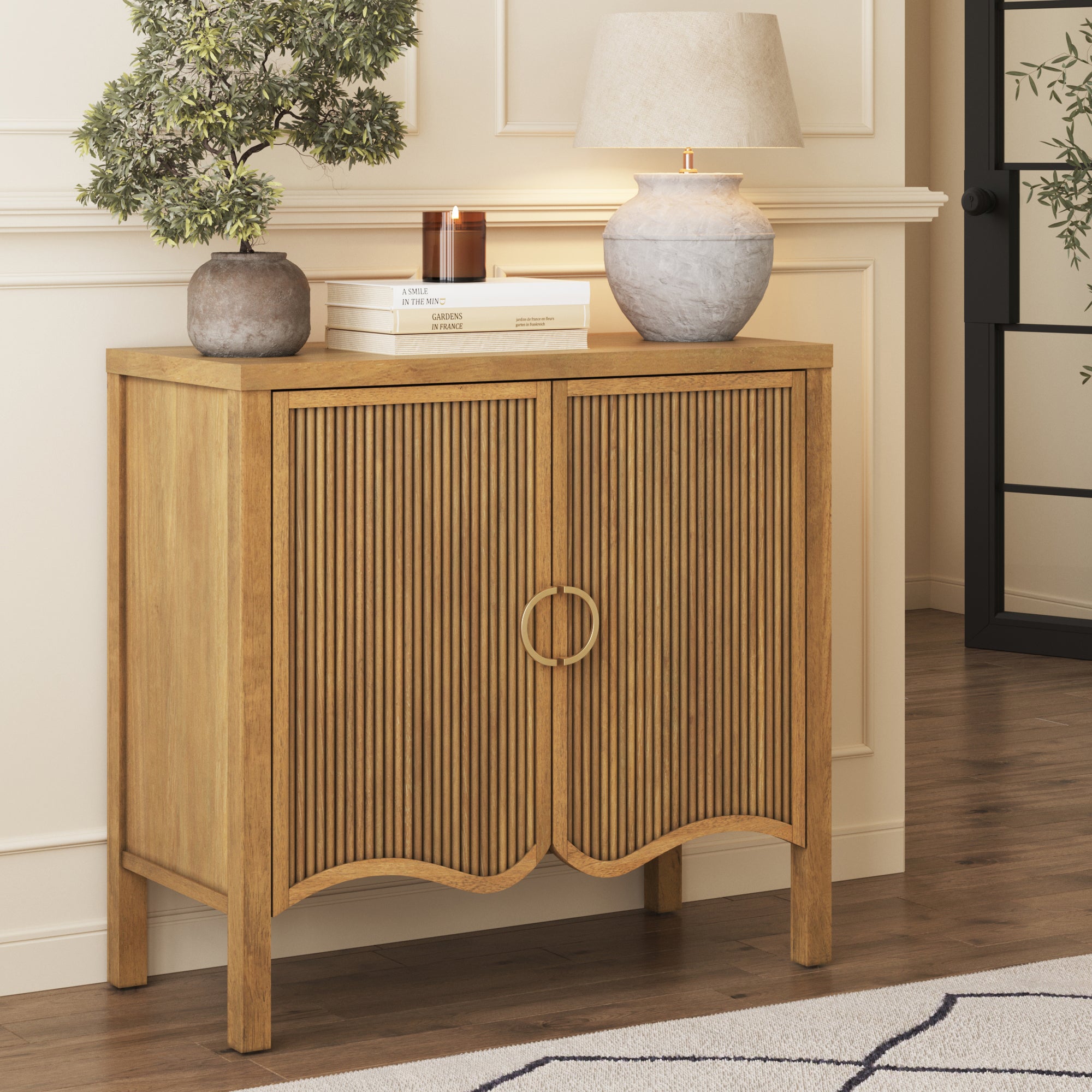 A Nathan James Transitional Fluted Cabinet with brass handle and solid wood legs stands against a cream wall, topped with a potted plant, books, candle, and beige ceramic lamp. A rug and wooden floor are partly visible.