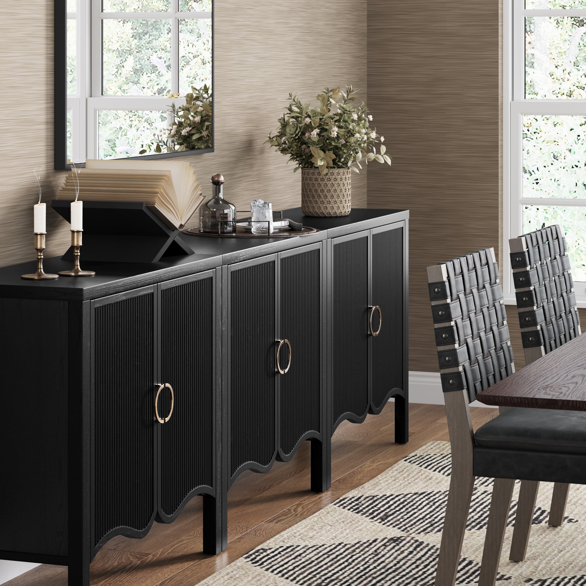 A modern dining room features the Nathan James Transitional Fluted Cabinet or TV Stand Black Oak (Set of 3) with gold handles, styled with candles, books, glass decanters, and a potted plant. A mirror, patterned rug, and wood table with woven chairs complete the space.