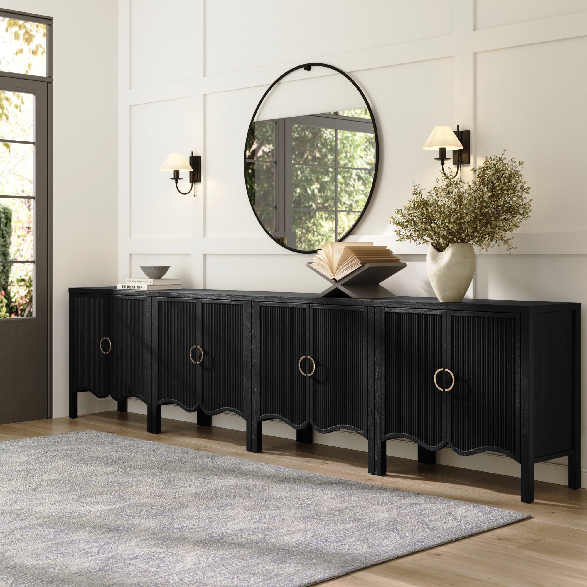 A modern hallway showcases a Nathan James Transitional Fluted Large Credenza Cabinet in black oak (set of 4), gold handles, a round mirror, two wall sconces, a vase with branches, an open book, light wood floors, and a gray rug.