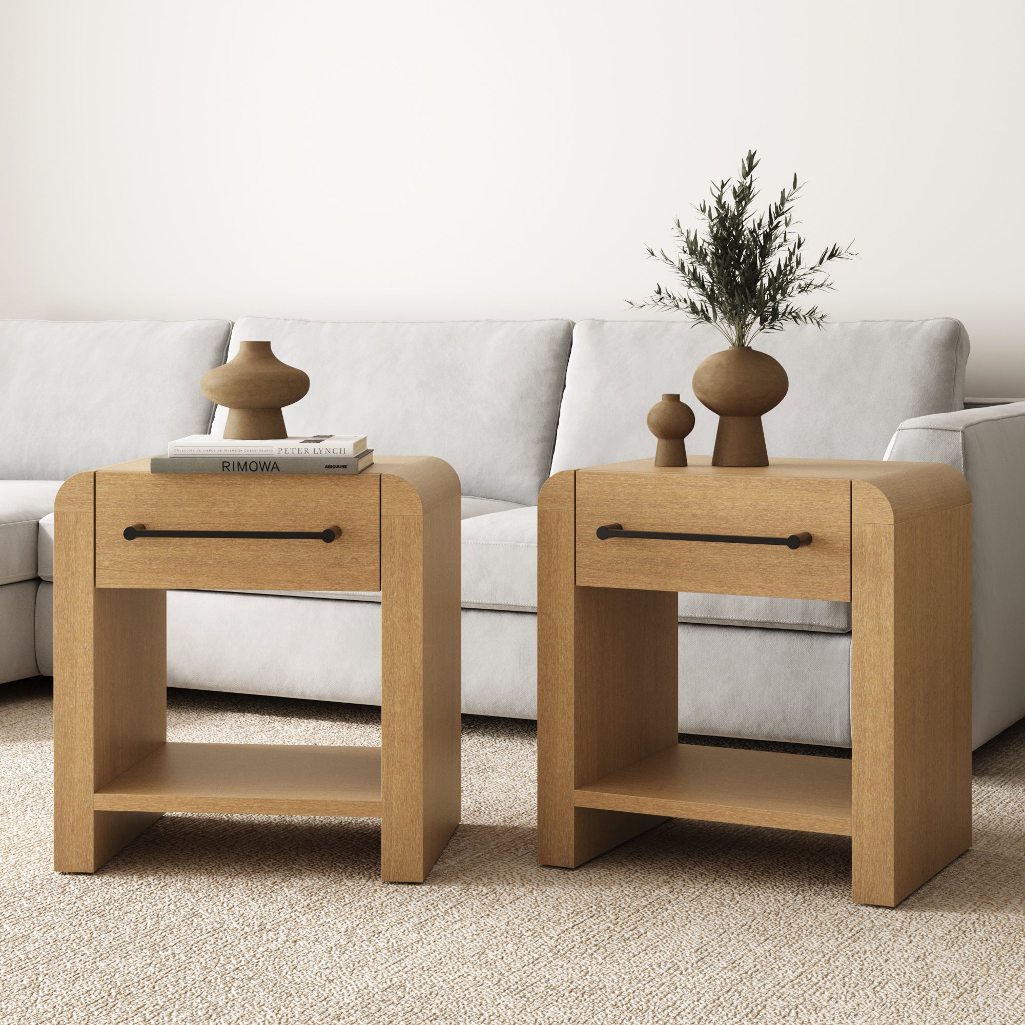 Two Nathan James Modern 1-Drawer Nightstands with metal handles in light brown sit before a light gray sofa—one holds stacked books and vases, the other displays decorative vases with greenery.