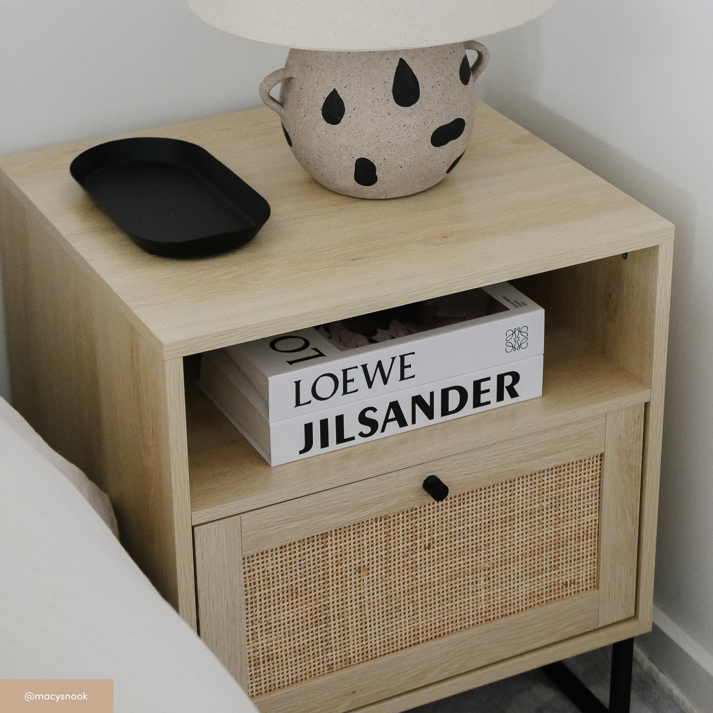 A light wood nightstand with a woven drawer front holds two books—one titled LOEWE and the other JILSANDER. On top are a speckled lamp and a black tray. The nightstand sits beside a bed.