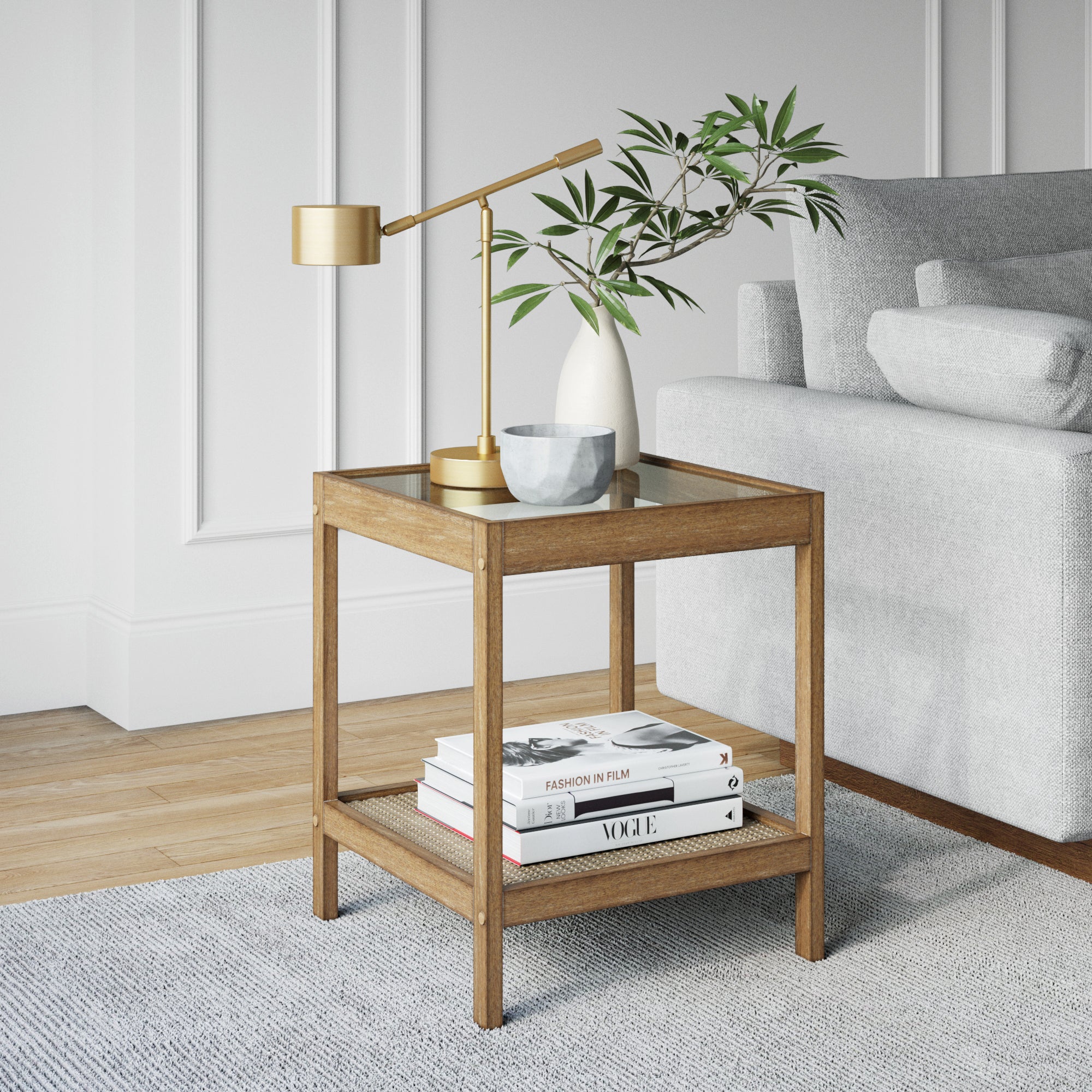 A wooden side table with a gold lamp, a small plant in a white vase, and a gray bowl on top. Four books are stacked on the lower shelf. The table sits next to a light gray sofa on a textured rug.
