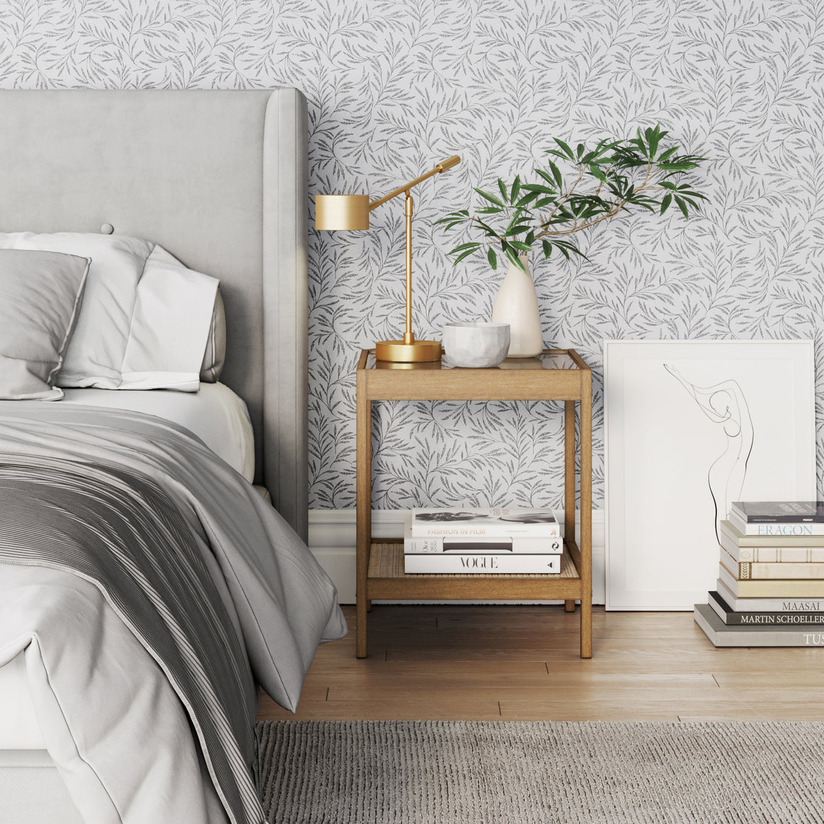 A modern bedroom features a gray upholstered bed with white and gray bedding, a wooden nightstand holding a gold lamp, decor vases with green branches, and stacked books beside a minimalist line art drawing.