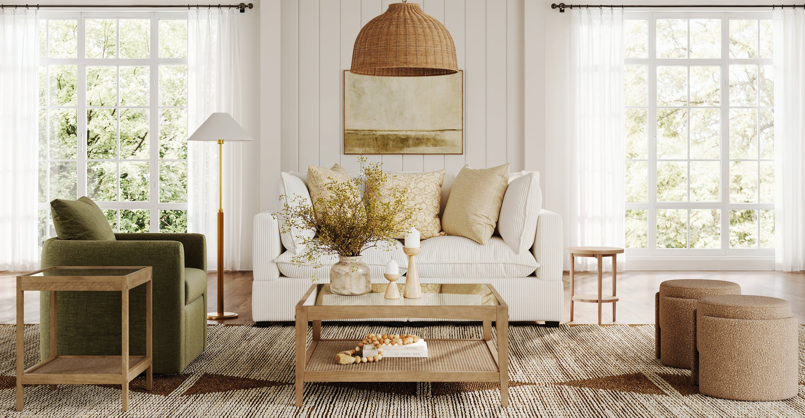 A bright living room with large windows, a white sofa with neutral cushions, a green armchair, wooden tables, woven stools, and natural decor including a rattan pendant light and a vase of branches on the coffee table.