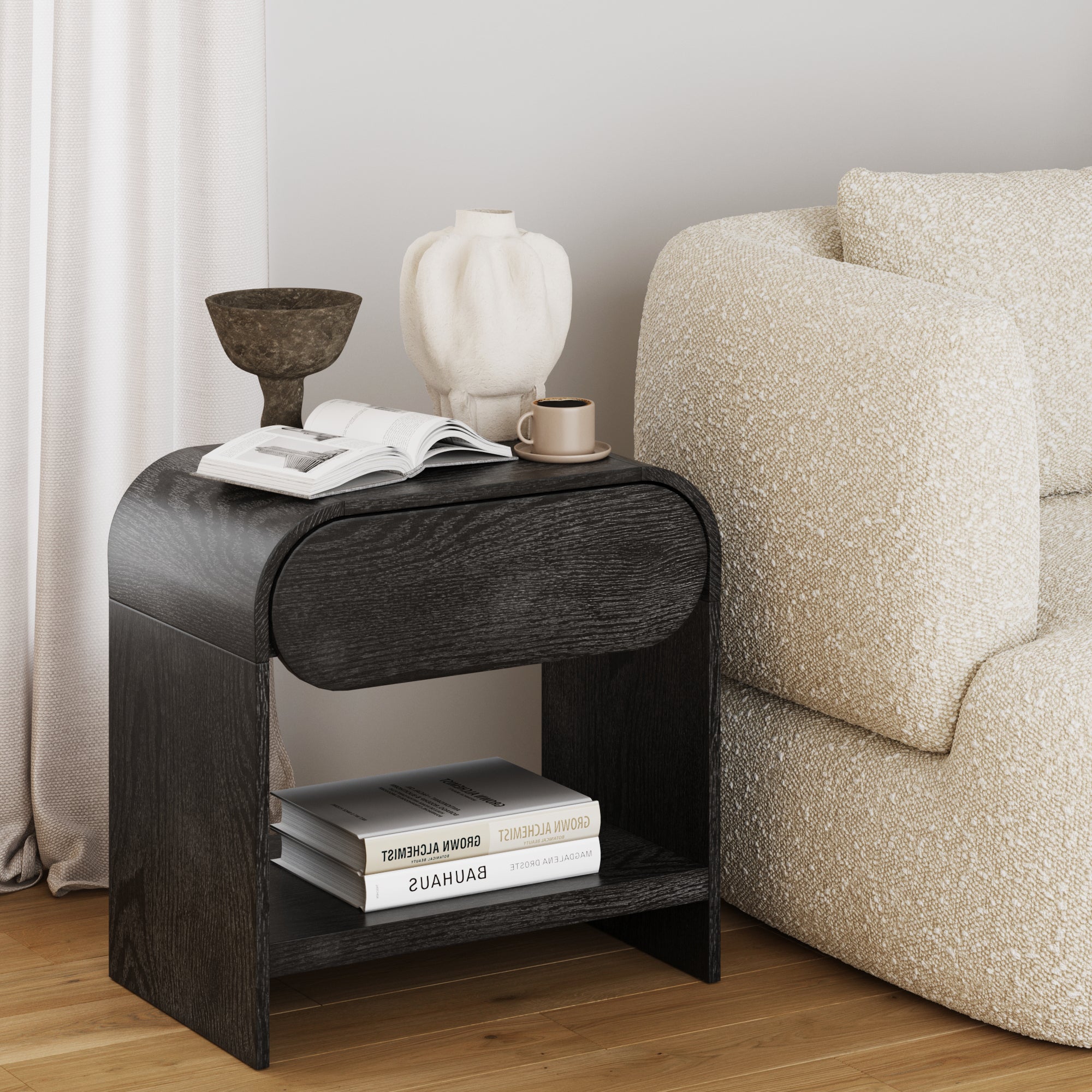 A modern black side table with a rounded drawer, holding an open book, a ceramic vase, a brown cup, and a decorative bowl, sits beside a textured cream sofa on a wooden floor.
