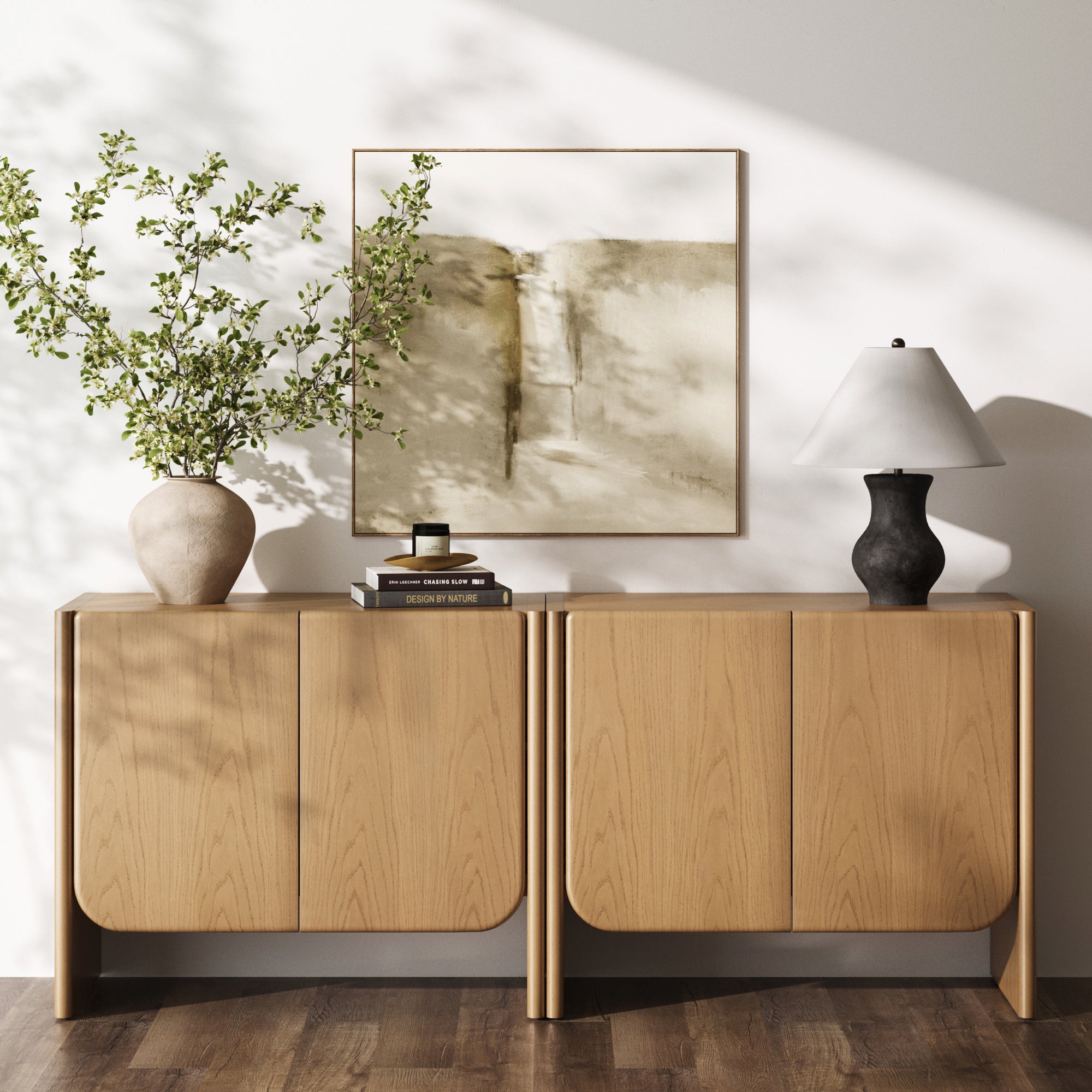 Two Nathan James Rounded Wood Accent Cabinets in Raw Oak stand side by side. On top are a beige vase with leafy branches, stacked books, a candle, and a black-base lamp with white shade. A neutral abstract painting hangs on the wall behind them.