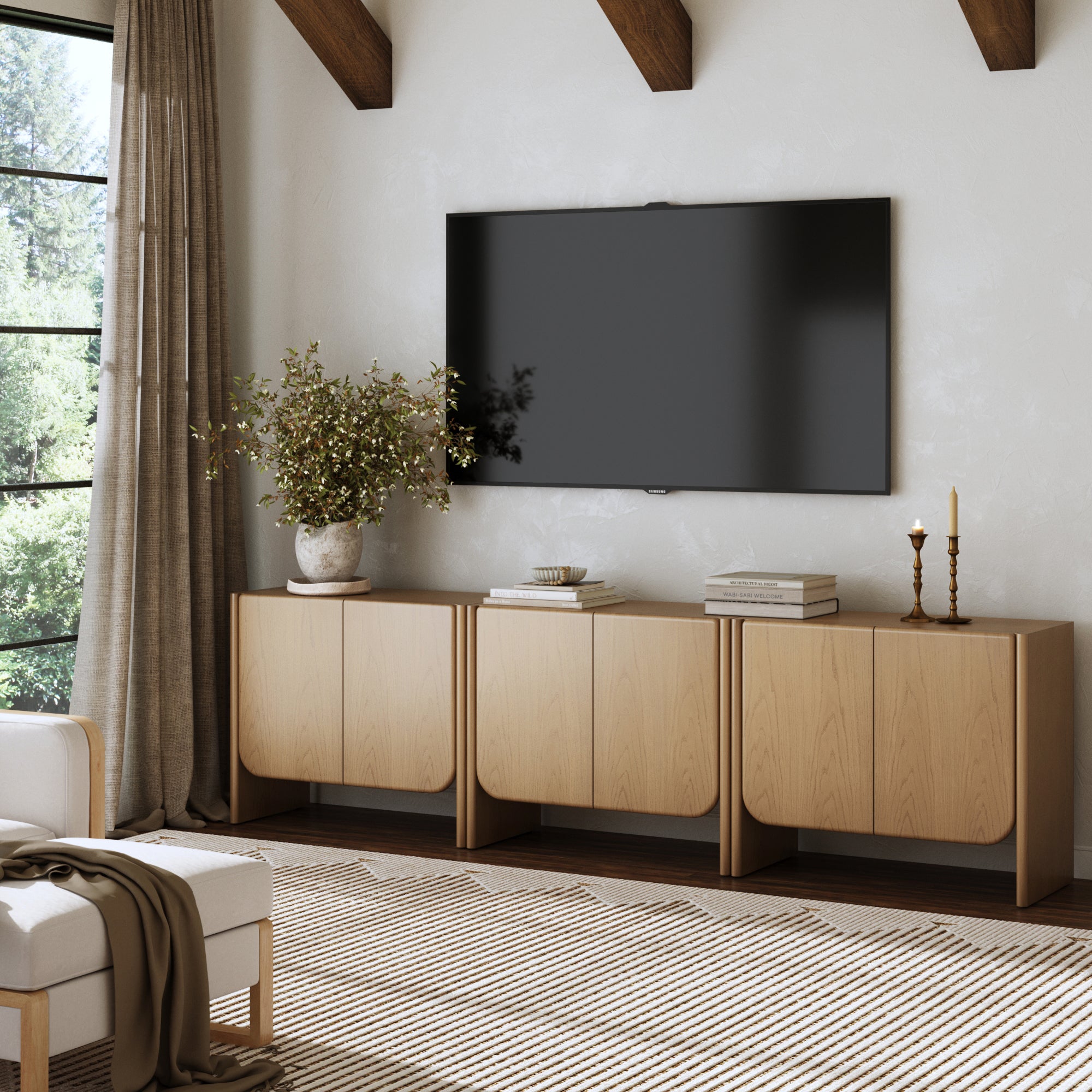 A modern living room featuring the Nathan James Rounded Wood Accent Cabinet or TV Stand in Raw Oak (Set of 3) beneath a wall-mounted TV, with a potted plant, stacked books, candles, a beige armchair, and brown curtains framing outdoor greenery.