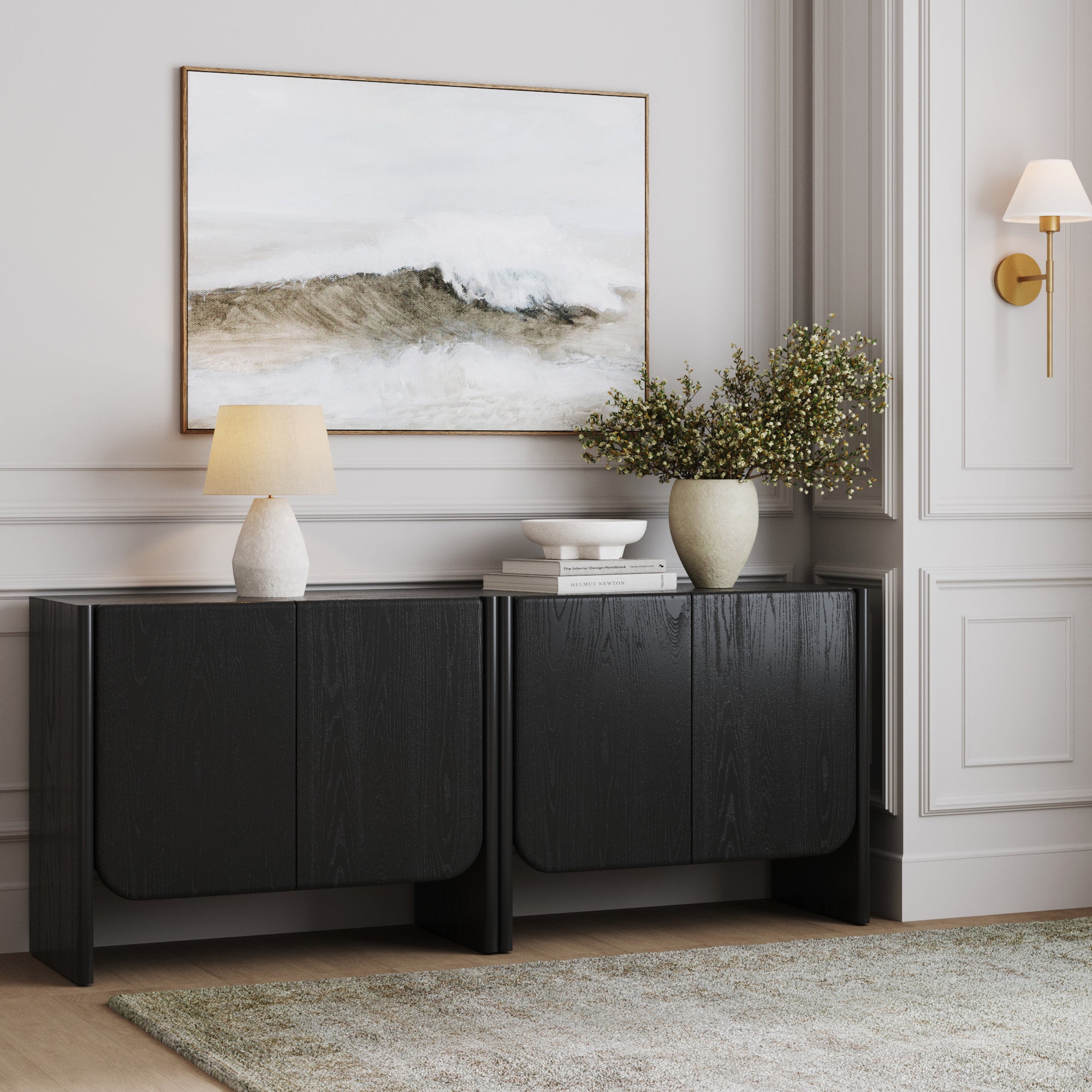 A modern interior features the Nathan James Rounded Wood Curio Cabinet or TV Stand in Black Oak (Set of 2), paired with a table lamp, green branches in a vase, a bowl, framed ocean wave art, gold wall sconce, light gray walls, and a soft rug.