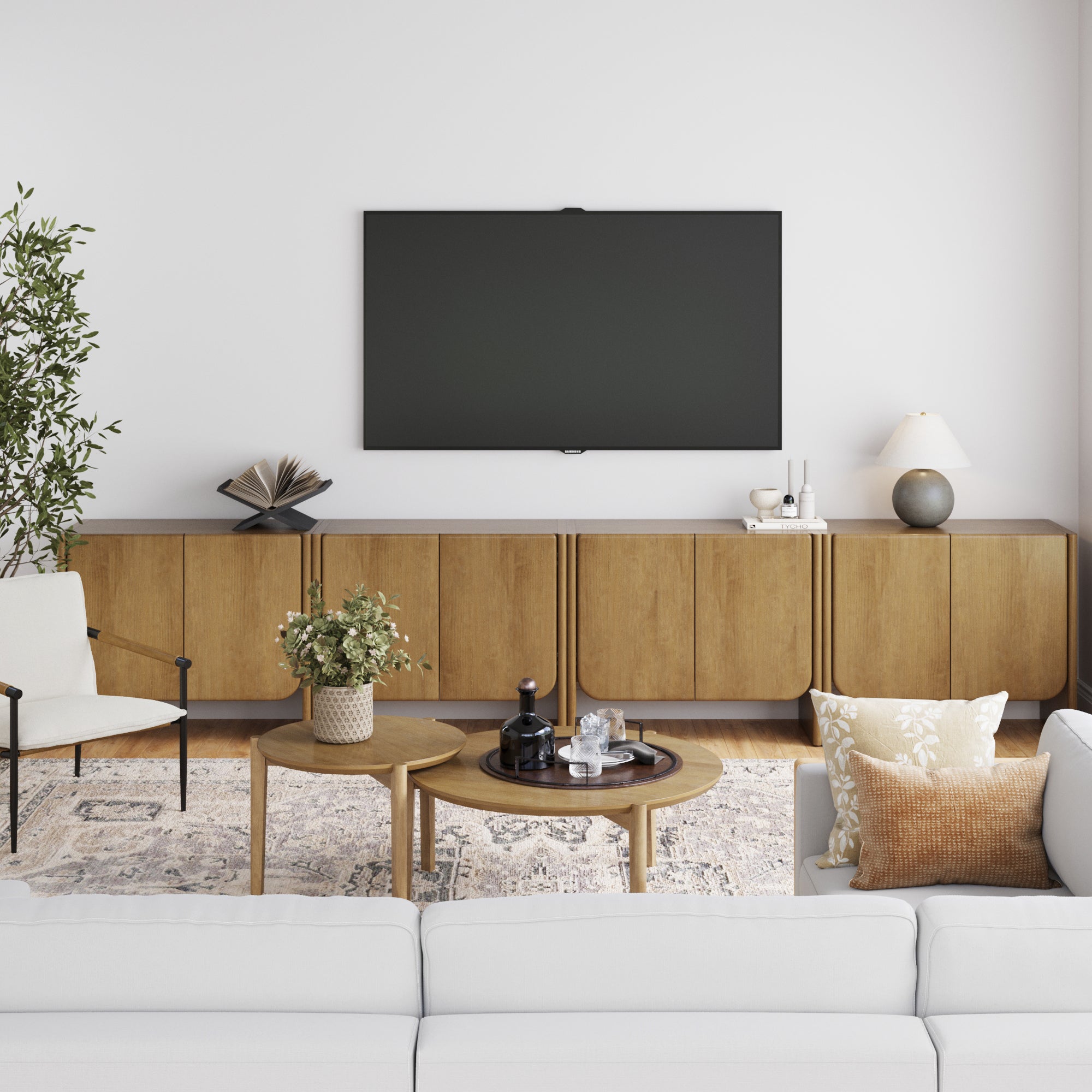 Modern living room featuring a wall-mounted TV, Nathan James Arched Wood Accent Cabinet or Large Credenza Light Brown (Set of 4), decorative items, potted plant, white armchair, round wooden coffee tables, and a white sofa with patterned cushions on a light rug.
