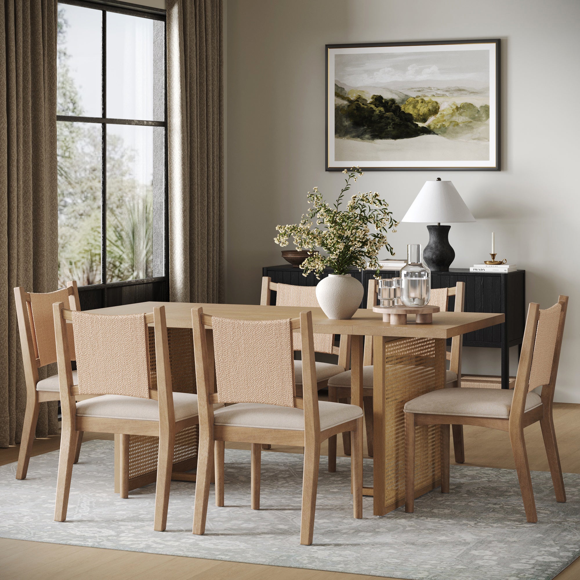 A modern dining room features the Nathan James Rattan Boho Dining Table with Seagrass Chairs (set of 6), a vase of greenery, glassware, and a sideboard with decor beneath a landscape painting. Large windows invite natural light.