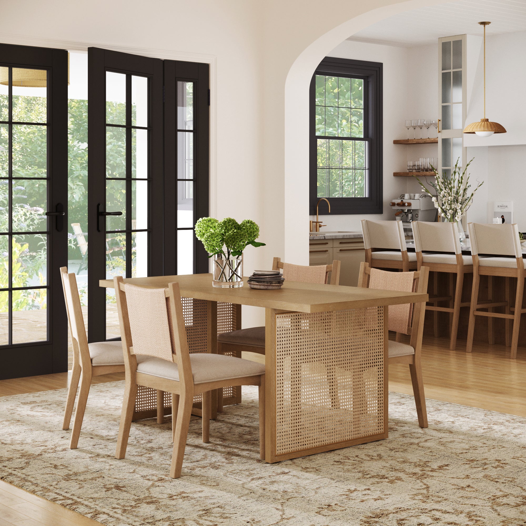 A modern dining room features the Nathan James Rattan Boho Dining Table with Seagrass Chairs Set of 4, a beige rug, and French doors to a green backyard. In the background, barstools, pendant light, and kitchen shelves are visible.