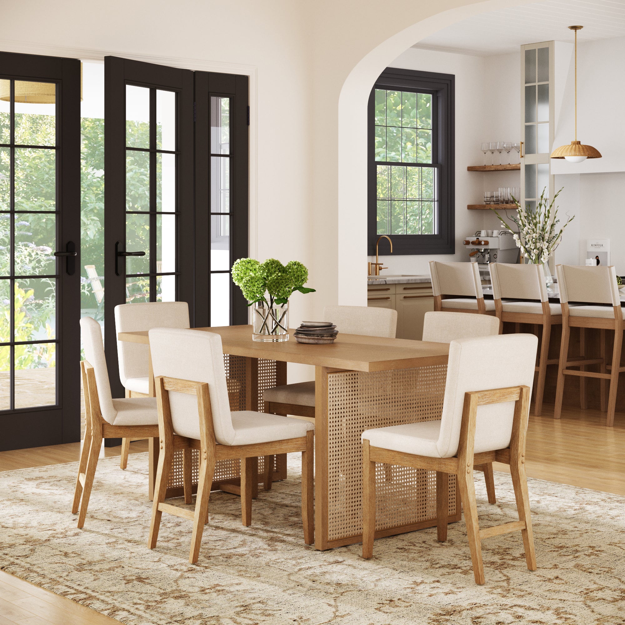 A modern dining room showcases the Nathan James Rattan Boho Dining Set with 6 upholstered chairs, a vase of green hydrangeas, black-framed windows, a neutral rug, and an open archway leading to a kitchen with pendant lighting.