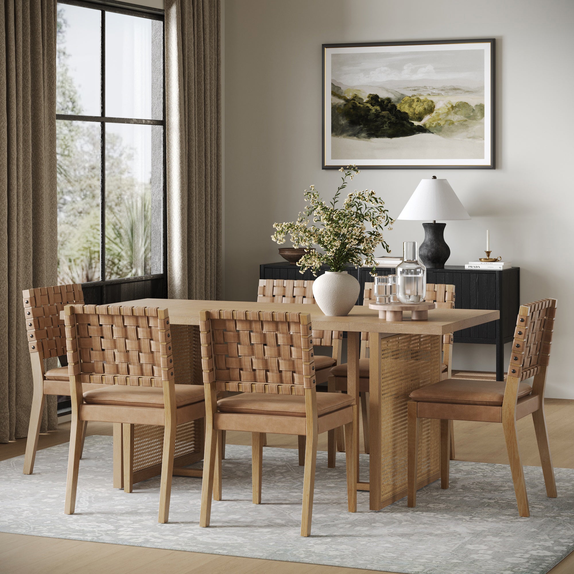 A modern dining room showcases the Nathan James Rattan Boho Dining Table Set of 6 with faux leather chairs, a large vase of greenery, and a framed landscape painting. Beige curtains filter light onto a patterned rug for an inviting atmosphere.