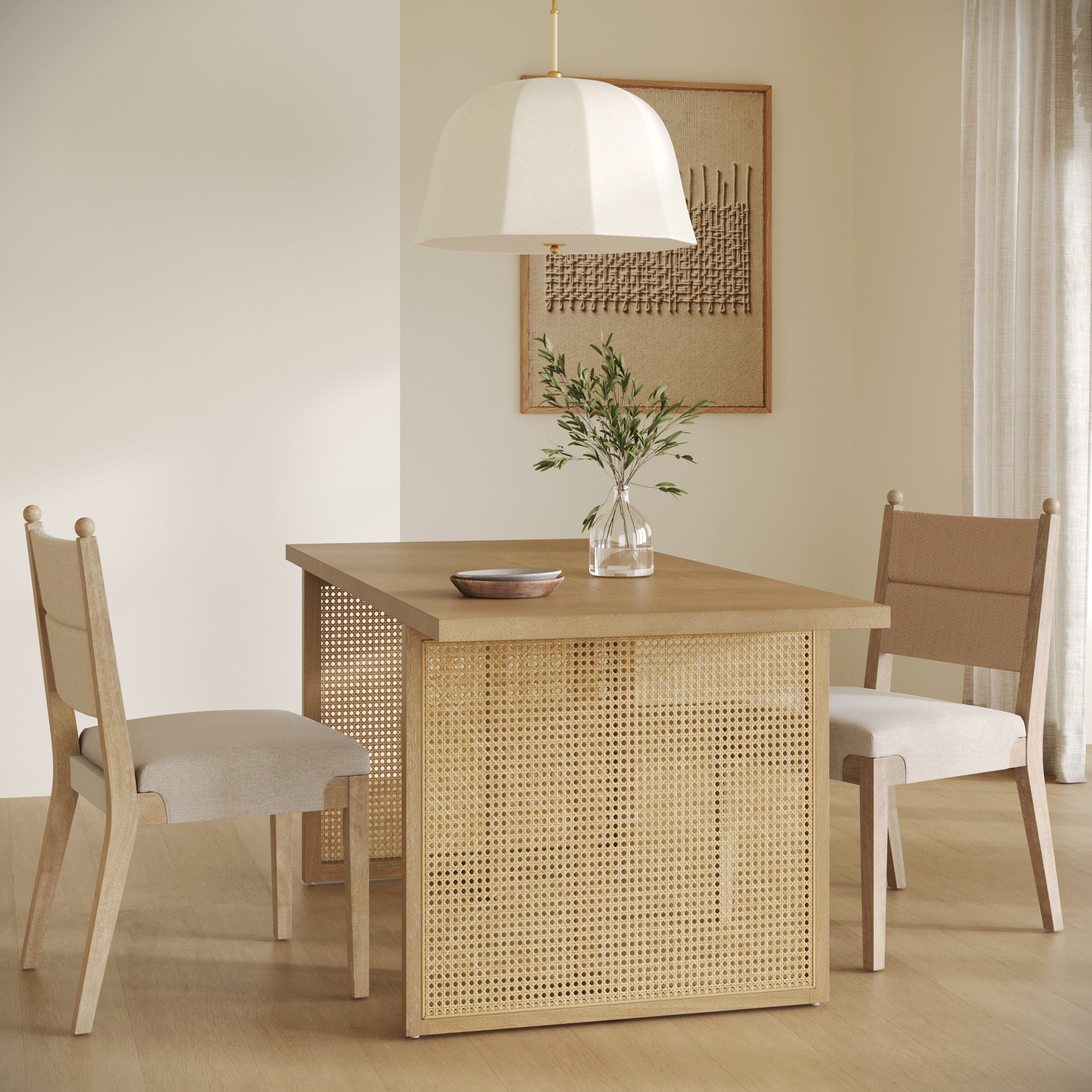 A minimalist dining area features the Nathan James Bohemian Natural Wood & Rattan Dining Table in light brown, two cushioned chairs, a pendant lamp, a vase with greenery, a bowl, and framed wall art—all in neutral tones with natural light.