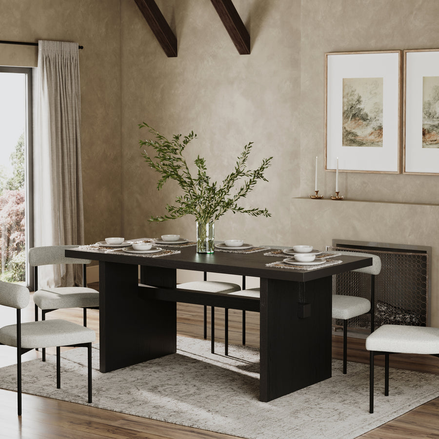 A modern dining room with the Nathan James Wood Modern Farmhouse Dining Table in Black Oak, six cushioned chairs, a leafy branch vase, neutral walls, framed art, candles on the mantel, and large windows with beige curtains.