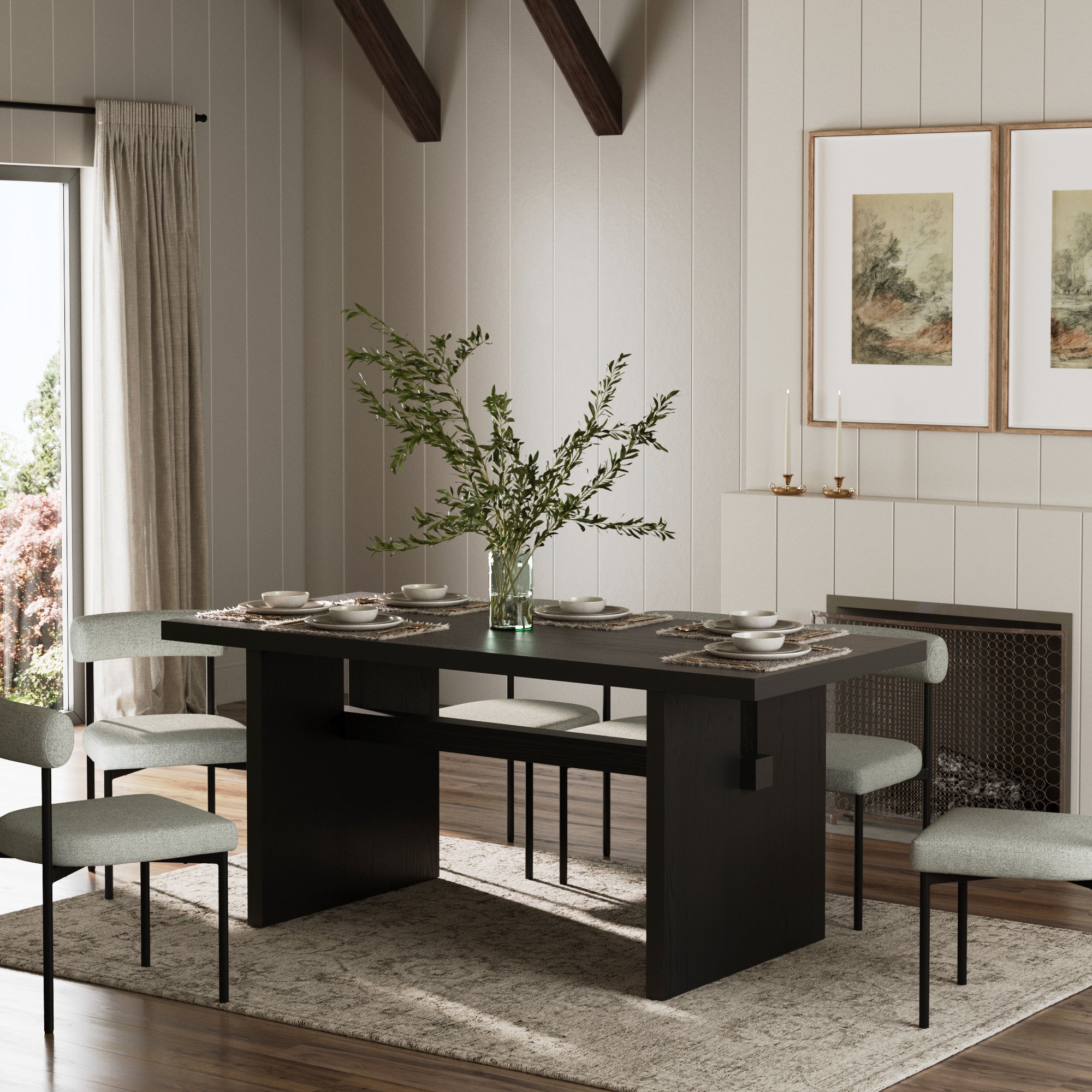 A modern dining room showcases the Nathan James Wood Modern Farmhouse Dining Table in black oak, seating 6 with upholstered chairs, a vase of green branches as centerpiece, neutral rug, two framed artworks, fireplace, and natural light from the left.