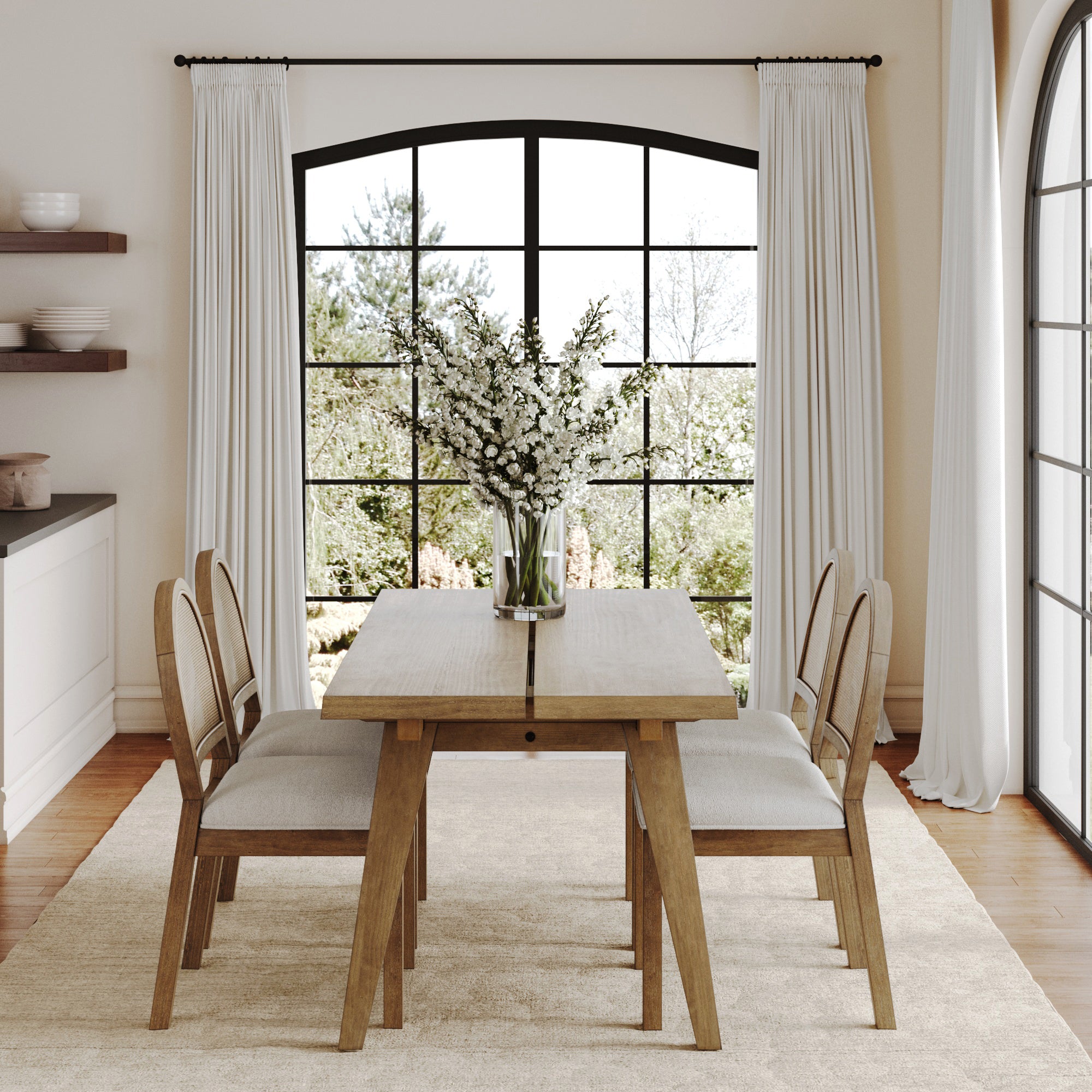 A bright dining room features the Nathan James Natural Rattan Dining Table Set with 4 chairs, a vase of white flowers, arched windows with white curtains, and shelves with dishes on the left. Sunlight enhances the room’s neutral palette.