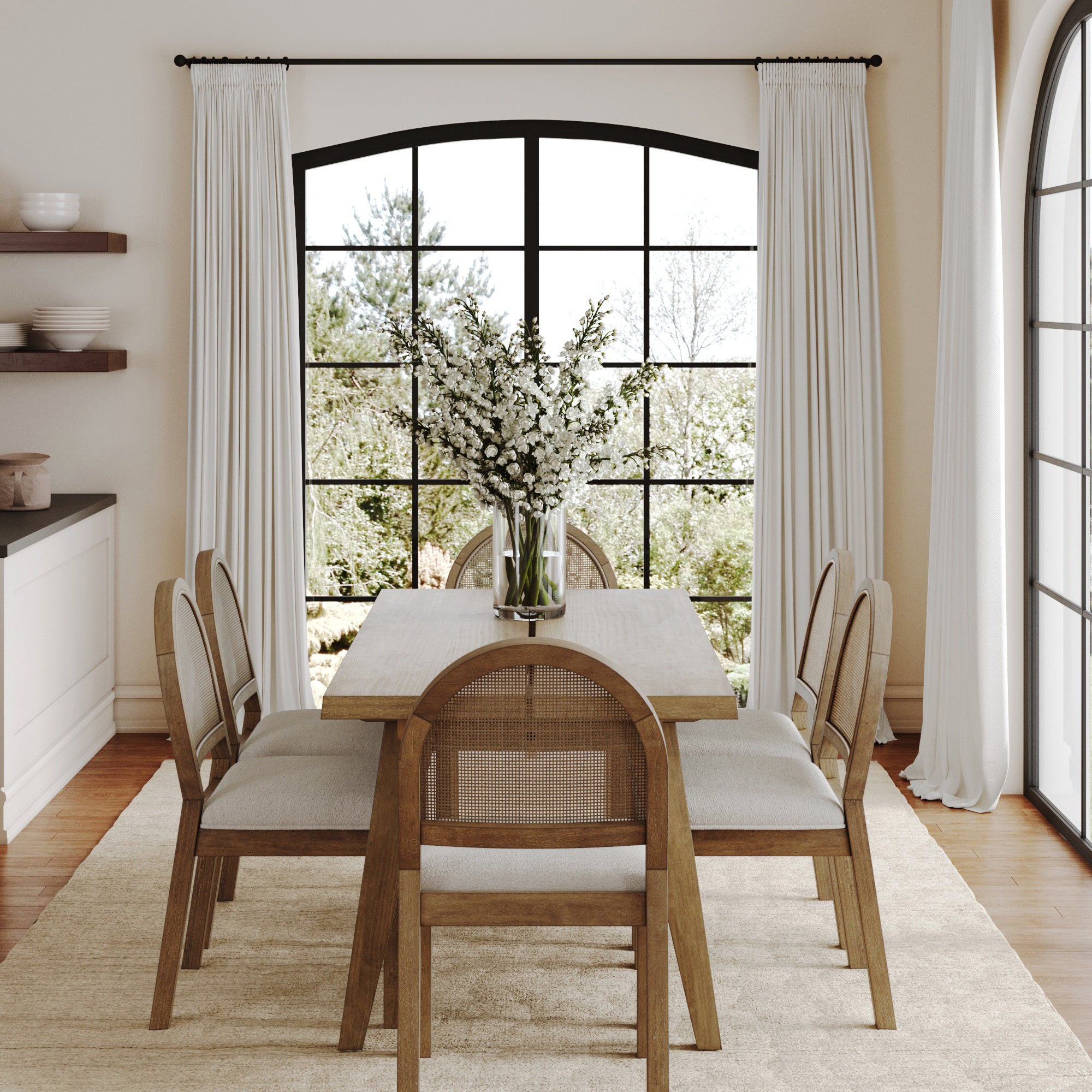A bright dining room features the Nathan James Natural Rattan Dining Table Set with six chairs, a vase of white flowers, arched windows with white curtains, a beige rug, and greenery visible outside.